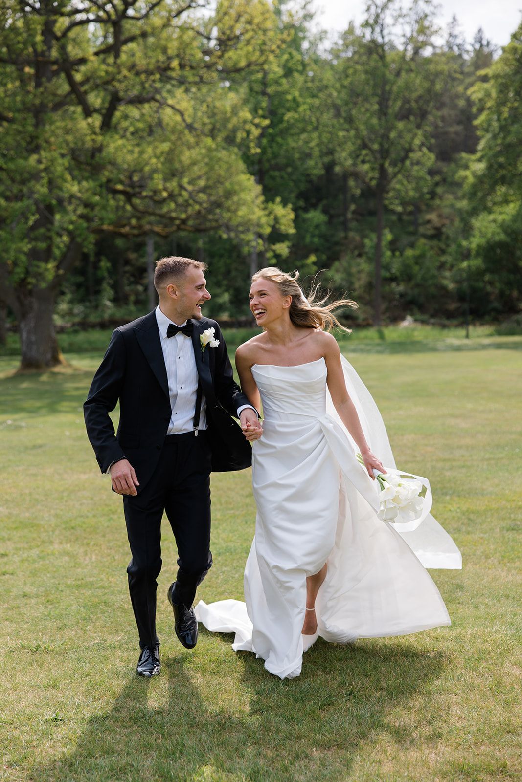 A bride and groom run playfully through a grassy field on their wedding day.