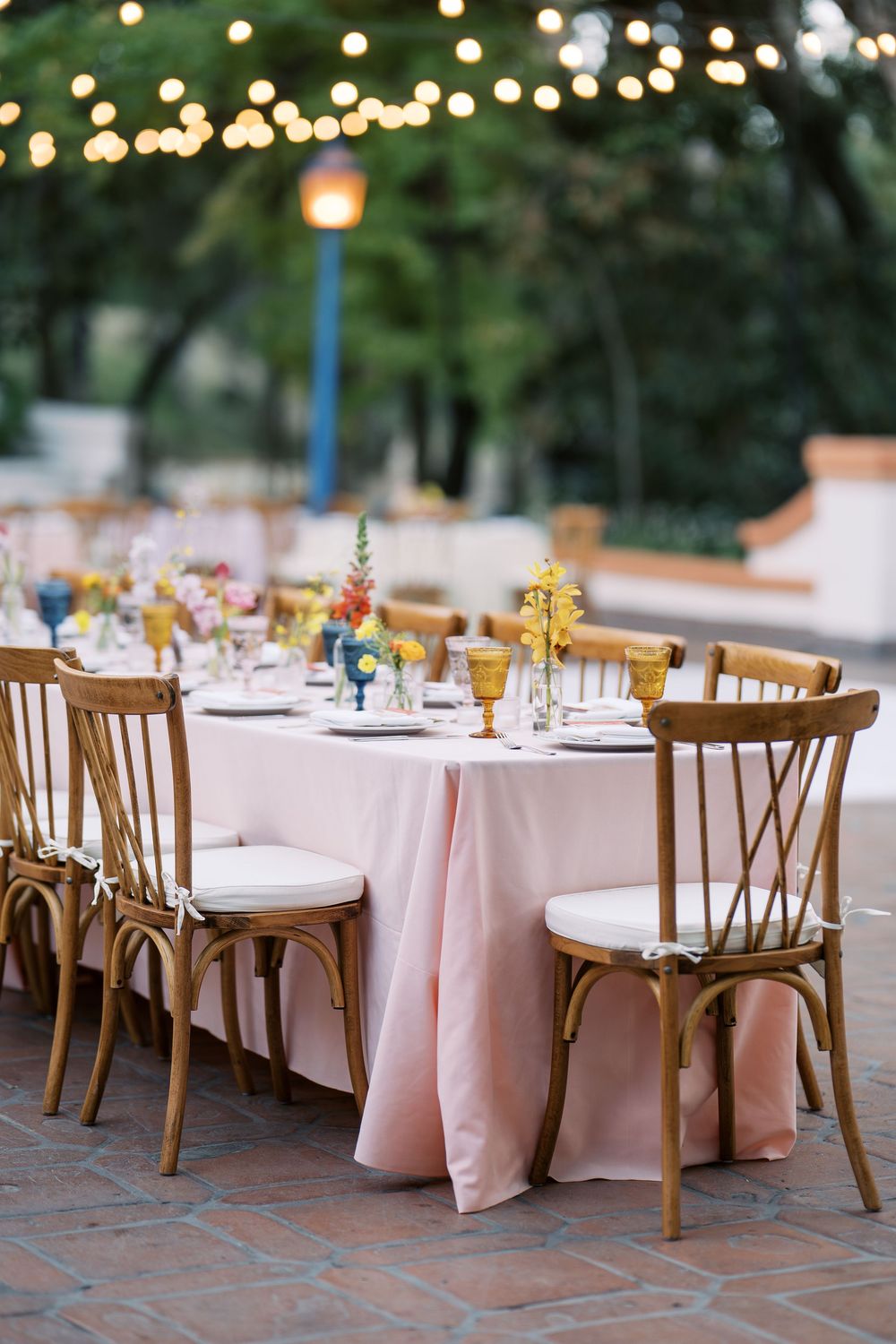 Outdoor wedding reception setup with wooden chairs and pink tablecloths under string lights at dusk.