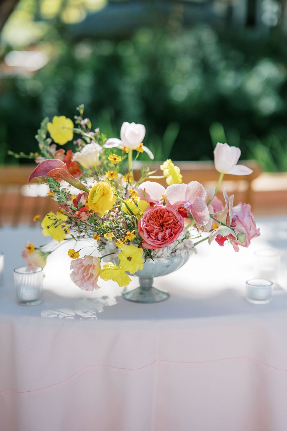 Colorful floral centerpieces with yellow, pink and coral flowers in glass vases arranged on a white tablecloth.