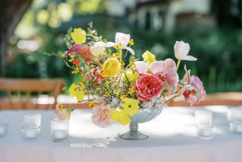 Elegant floral centerpieces with yellow and pink flowers in glass vases line a wedding reception table.