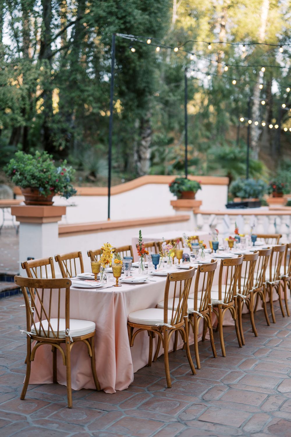 Long banquet tables with pink tablecloths and wooden chairs set for an elegant outdoor wedding reception under string lights.