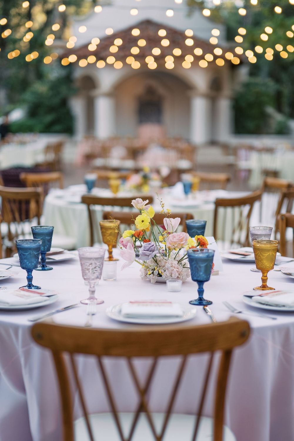 Elegant table settings with colorful vintage glassware in blue, amber and pink hues on white tablecloths at a formal event.