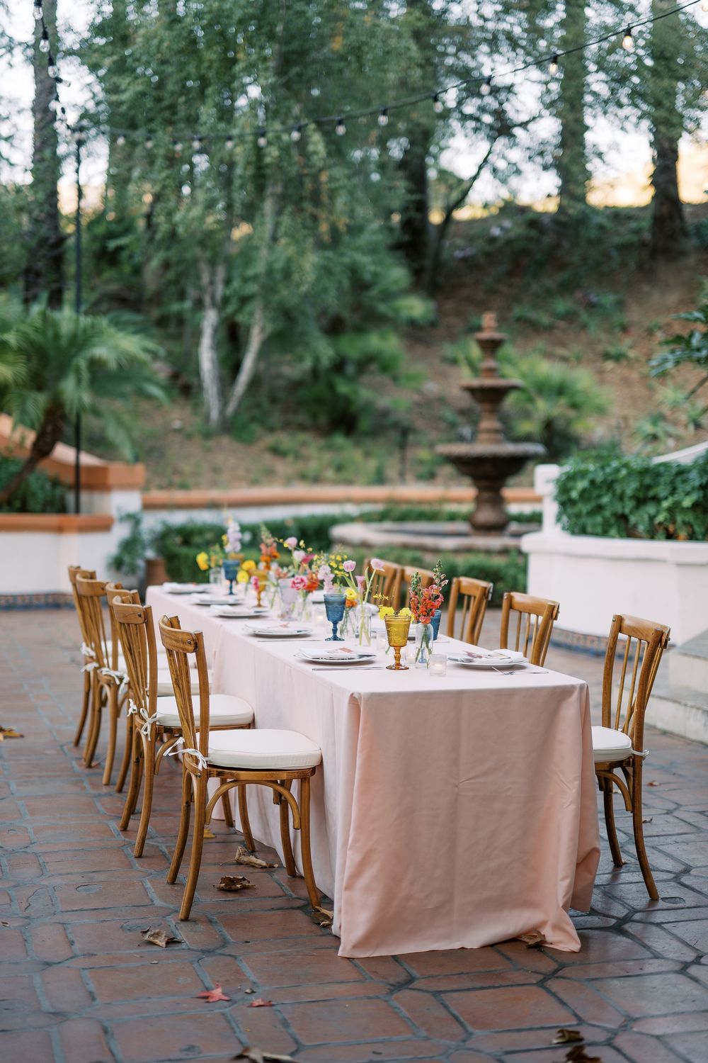 Round tables with blush pink linens and wooden chairs are elegantly arranged on a brick patio for an outdoor event.