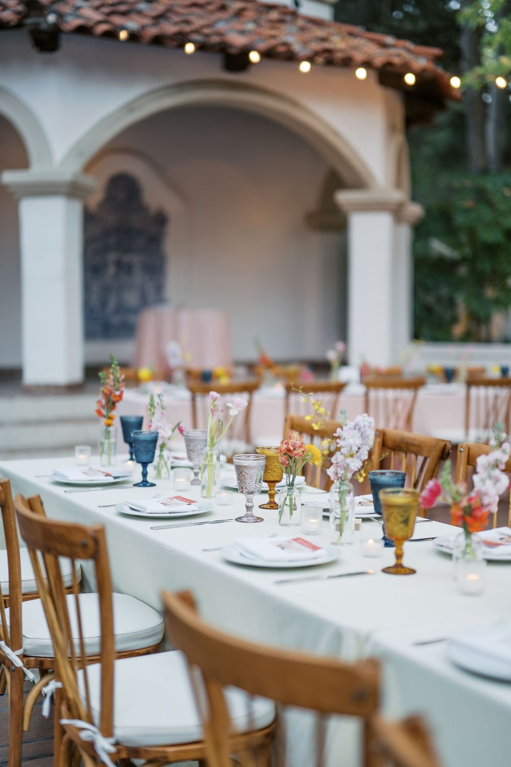 Elegant wedding reception table settings with wooden chairs and white linens under arched columns.