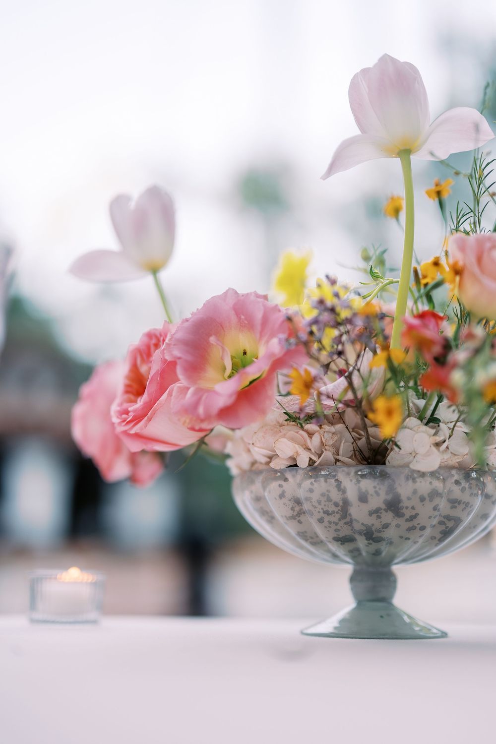 Delicate pink and white flowers arranged in an elegant glass pedestal bowl with soft dreamy bokeh background.