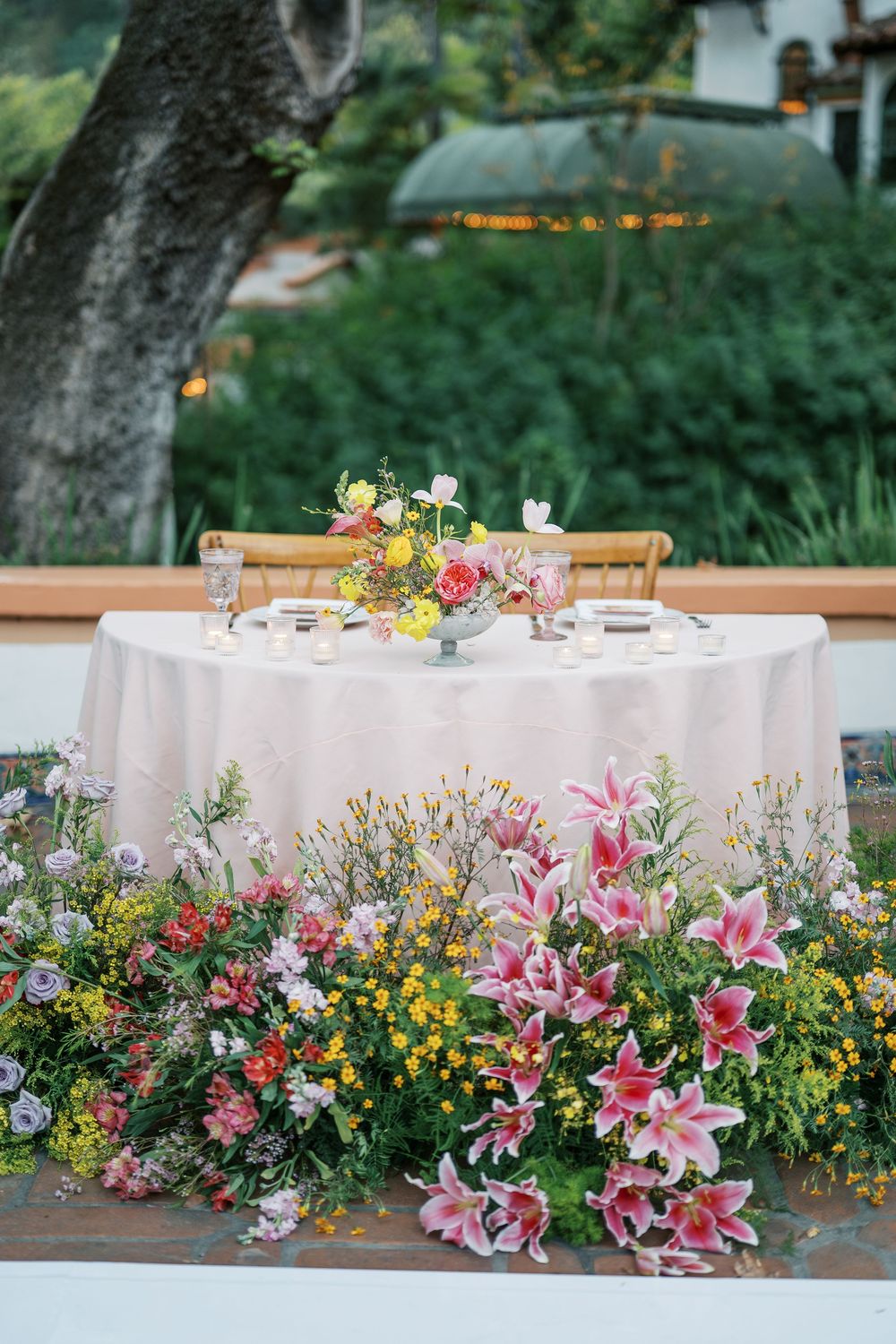 An elegant wedding table setting with white tablecloth surrounded by vibrant pink lilies and colorful wildflowers outdoors.