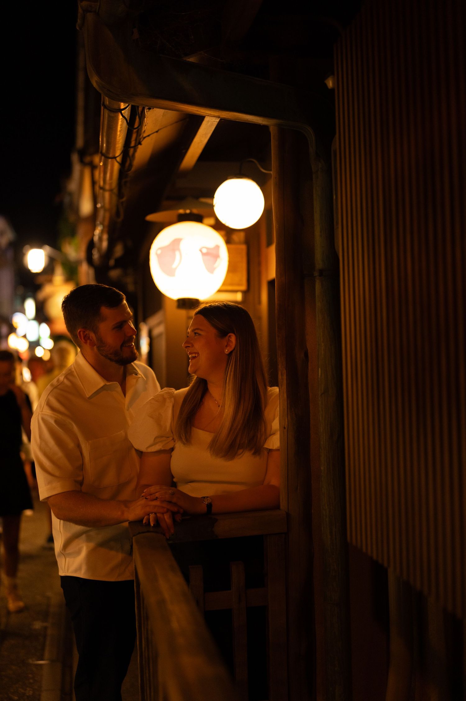 Couple laughing and holding hands outside a glowing Japanese lantern-lit street.