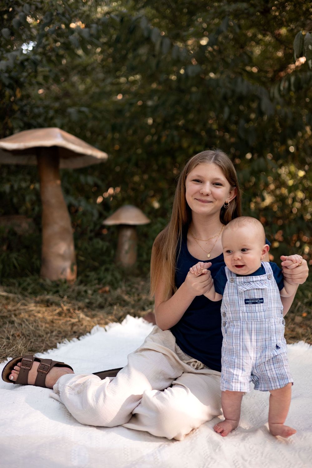 A brother and sister sit on a blanket in front of some giant mushrooms