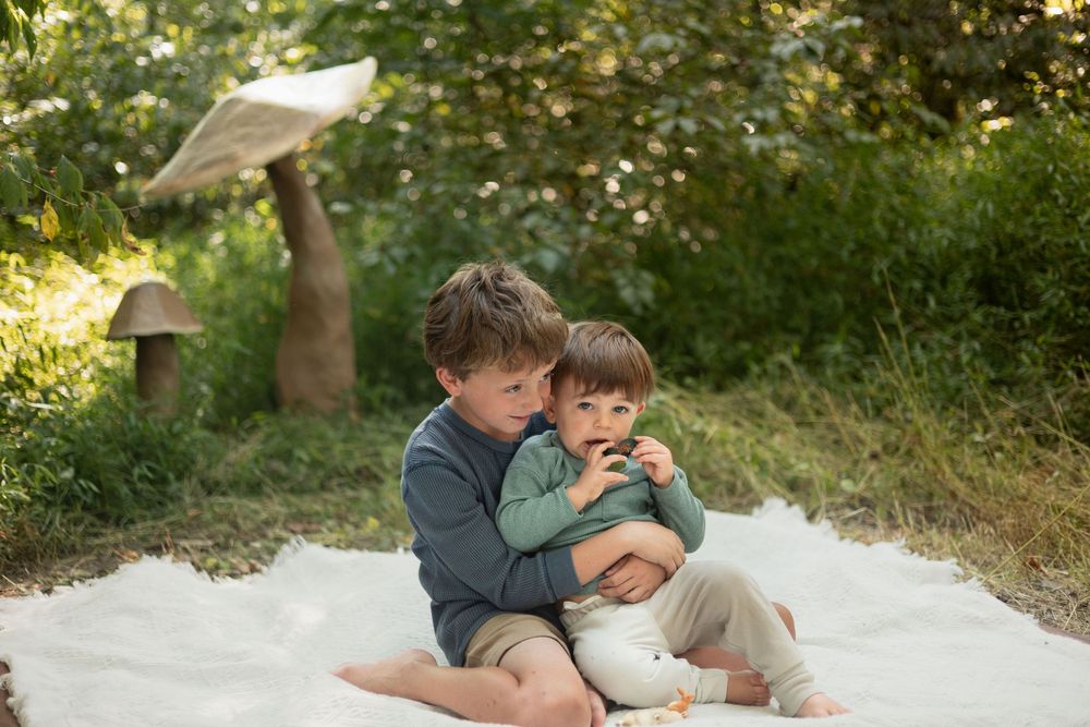 2 boys snuggle on a blanket with a giant mushroom in the back.
