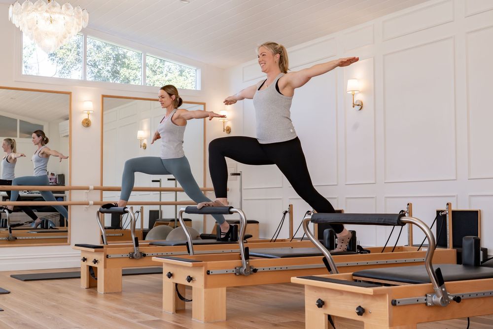 Pilates students performing synchronized movements on reformer equipment in an elegant white-walled fitness studio.