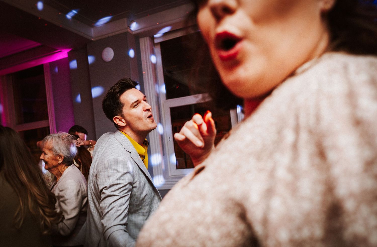 Party guests dancing and singing enthusiastically under purple lighting at an indoor celebration.