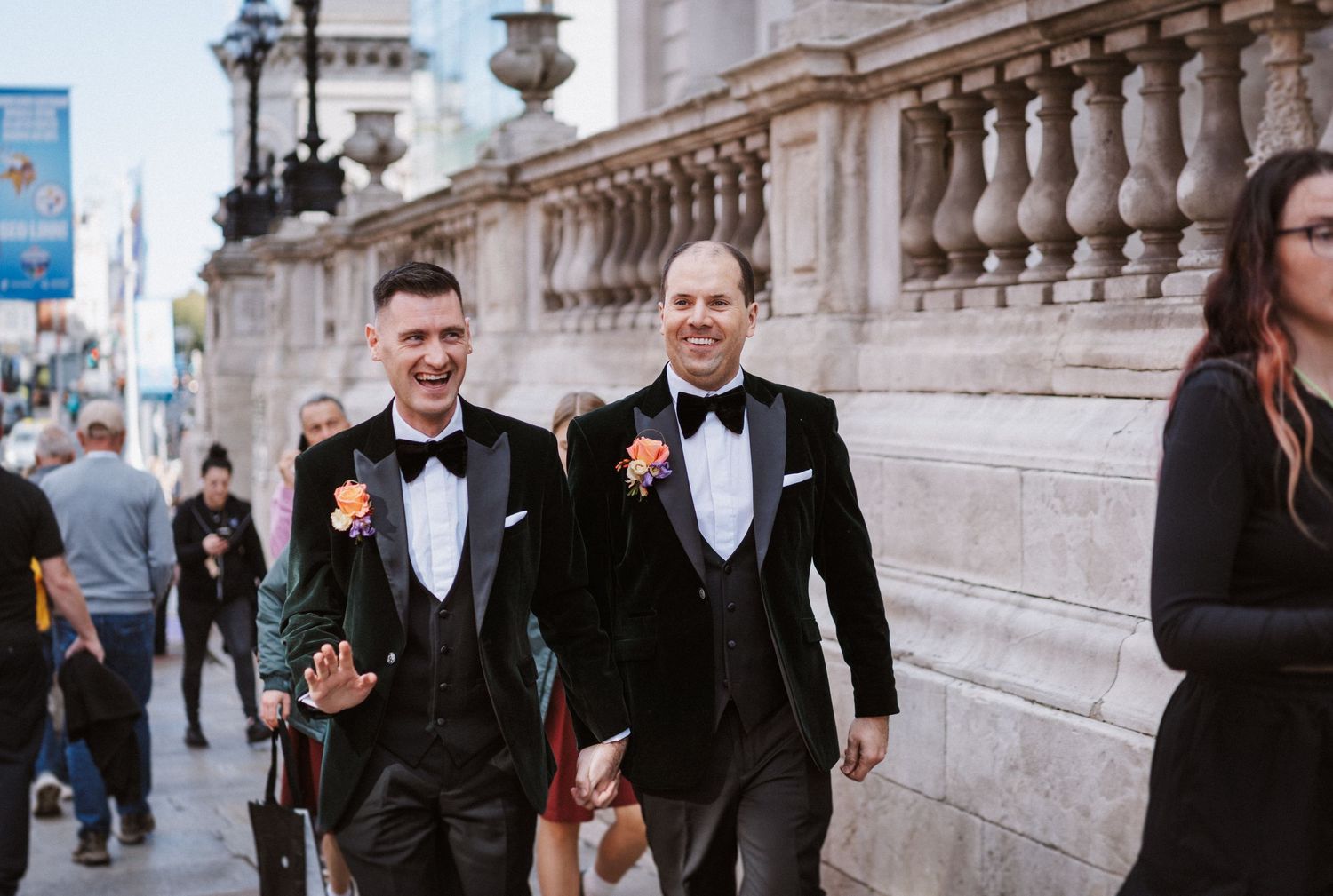 Groomsmen in black tuxedos walking together along an elegant building exterior.