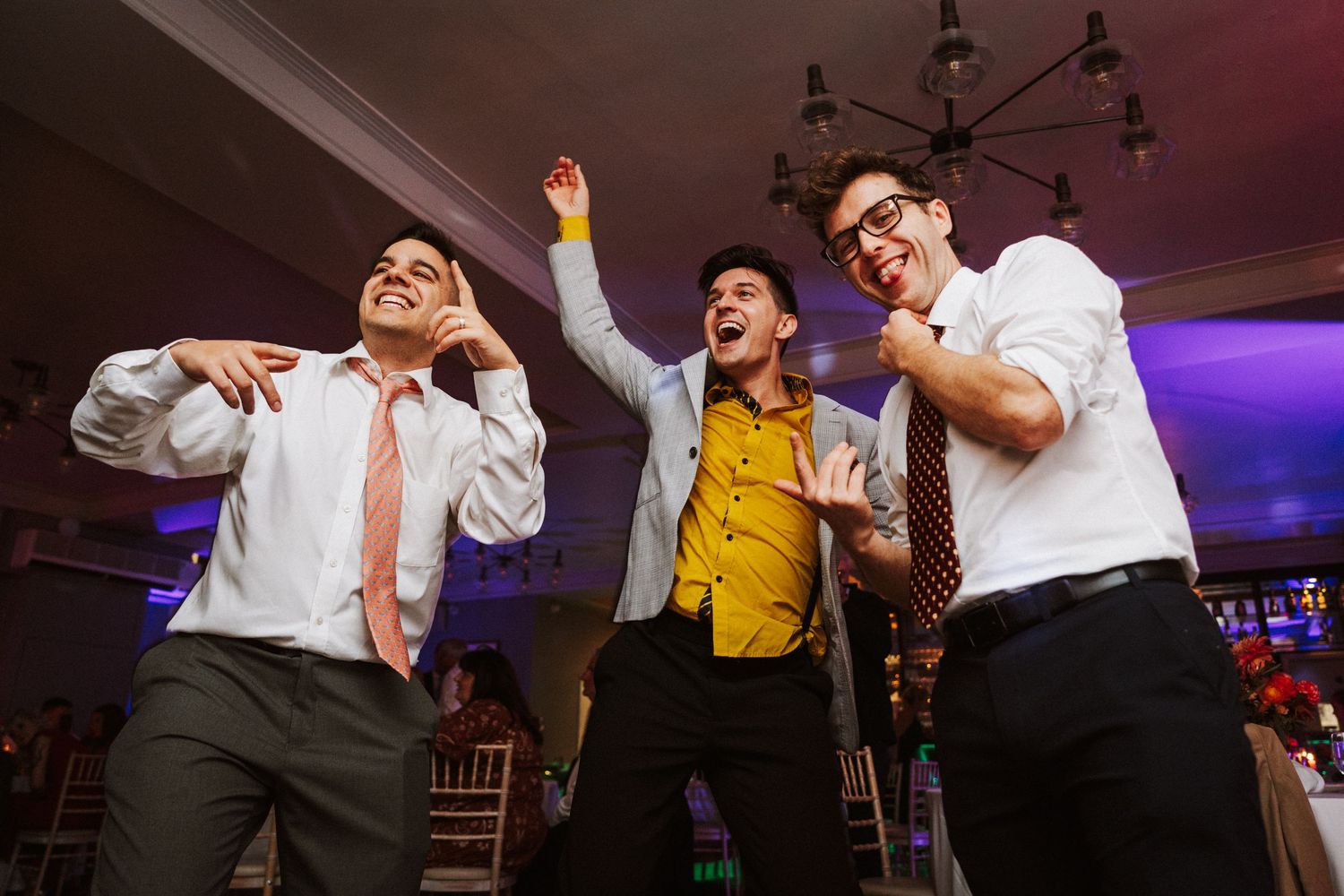 Wedding guests dancing enthusiastically under colorful lighting at an indoor reception venue.