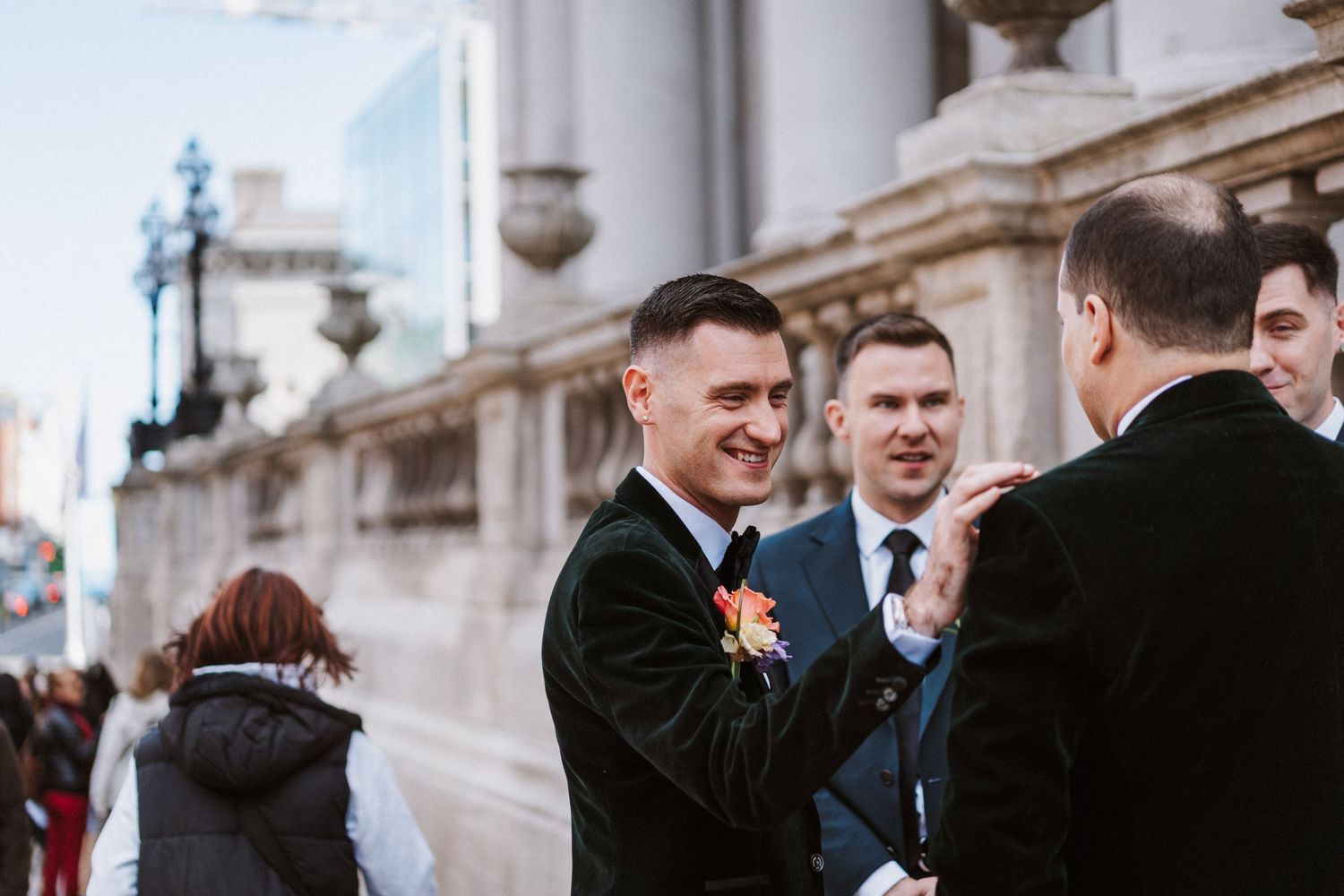 Wedding guests socialize in front of an elegant neoclassical building with stone columns.
