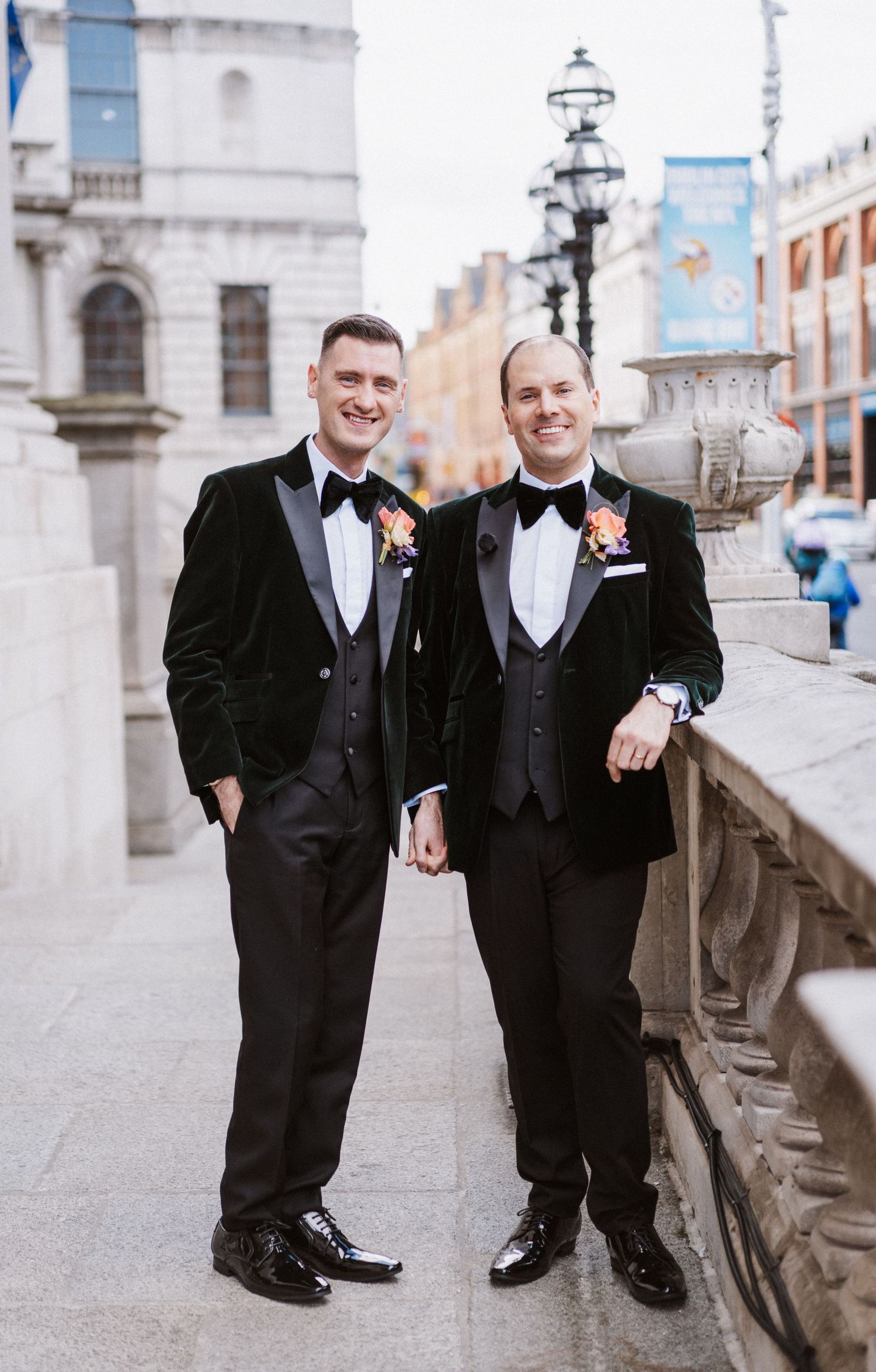 Two people in black tuxedos posing together along an outdoor stone walkway with vintage lampposts.