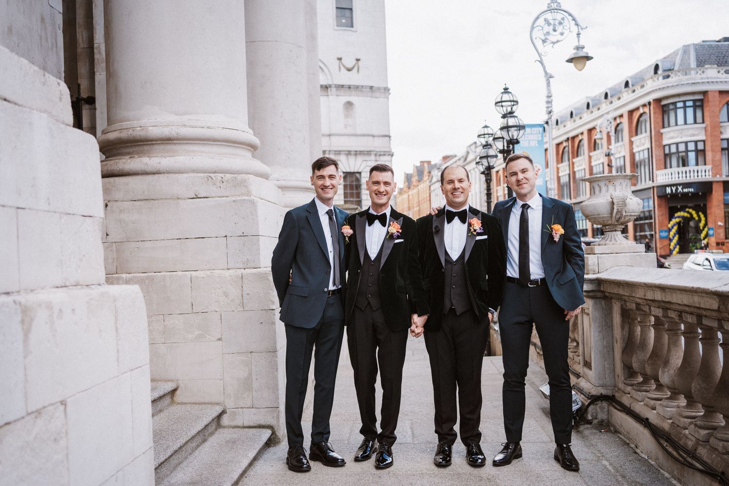 Wedding party in formal attire poses between classical white columns of historic building.