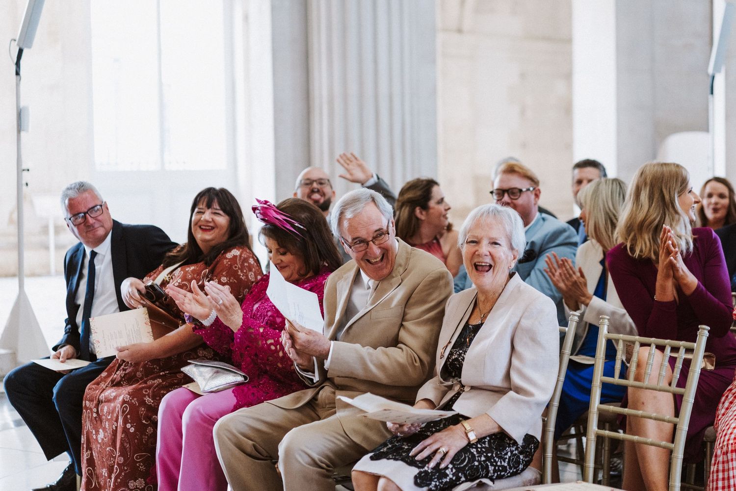 Wedding guests seated in rows during an indoor ceremony with natural lighting streaming through windows.