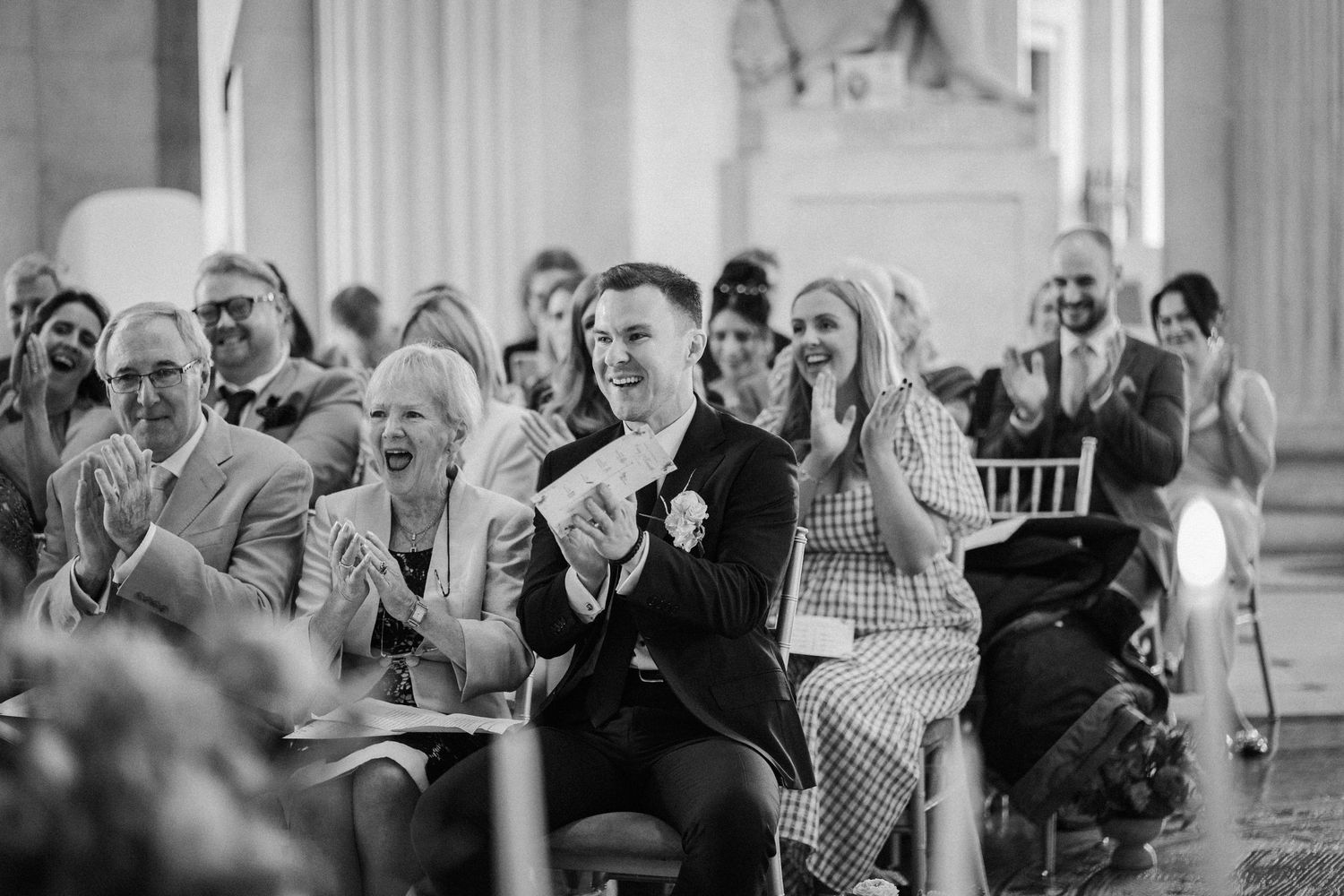 Wedding guests react with joy and laughter during an emotional moment at an elegant indoor ceremony.