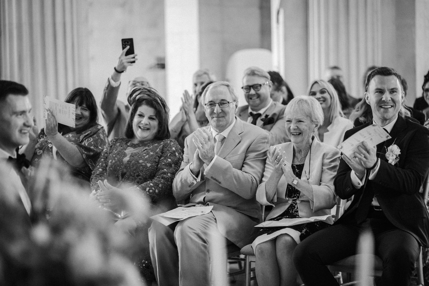 Wedding guests seated in white chairs during an indoor ceremony with classical columns.