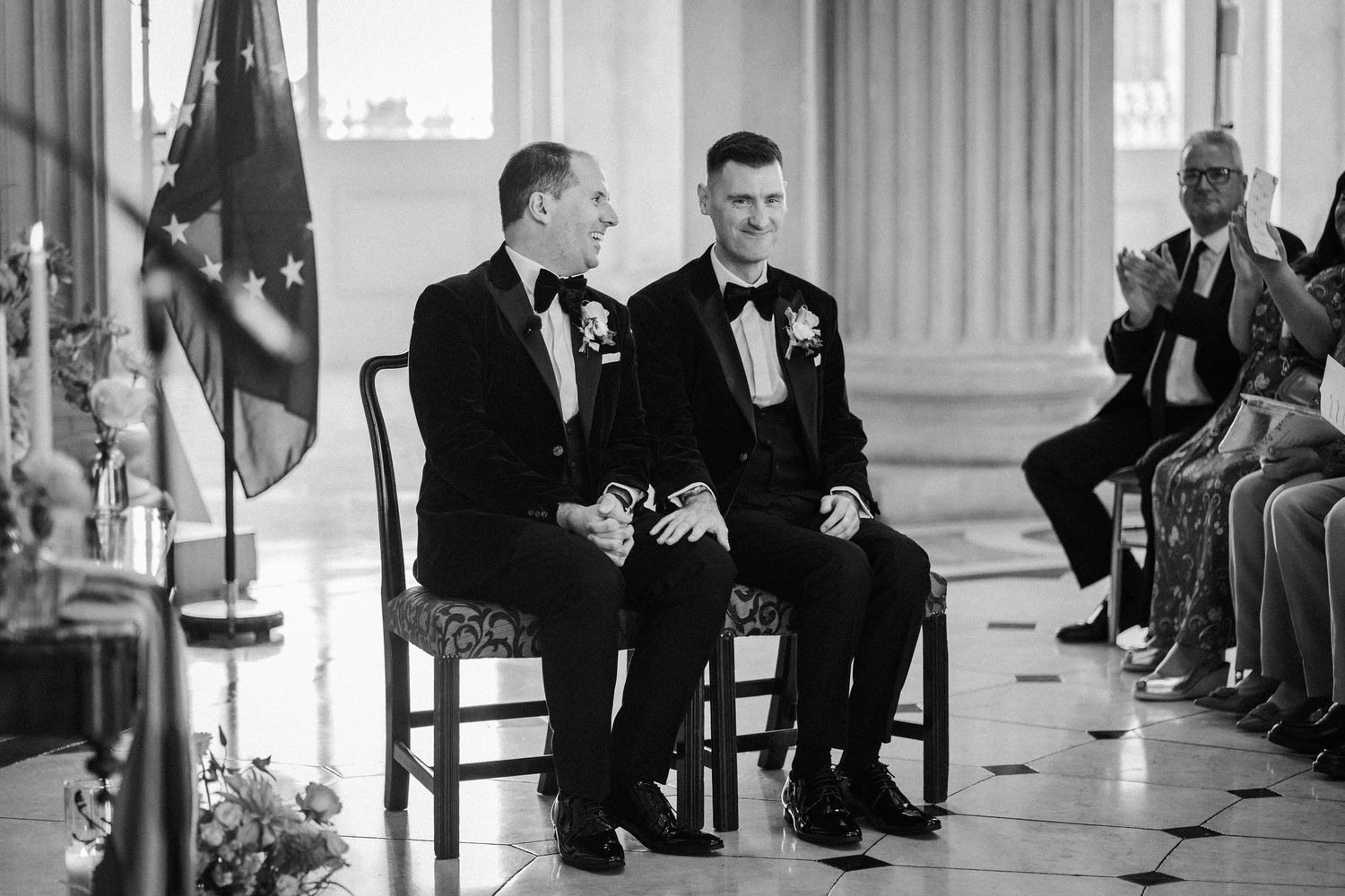 Wedding ceremony attendees gathered in an elegant columned venue with natural lighting.