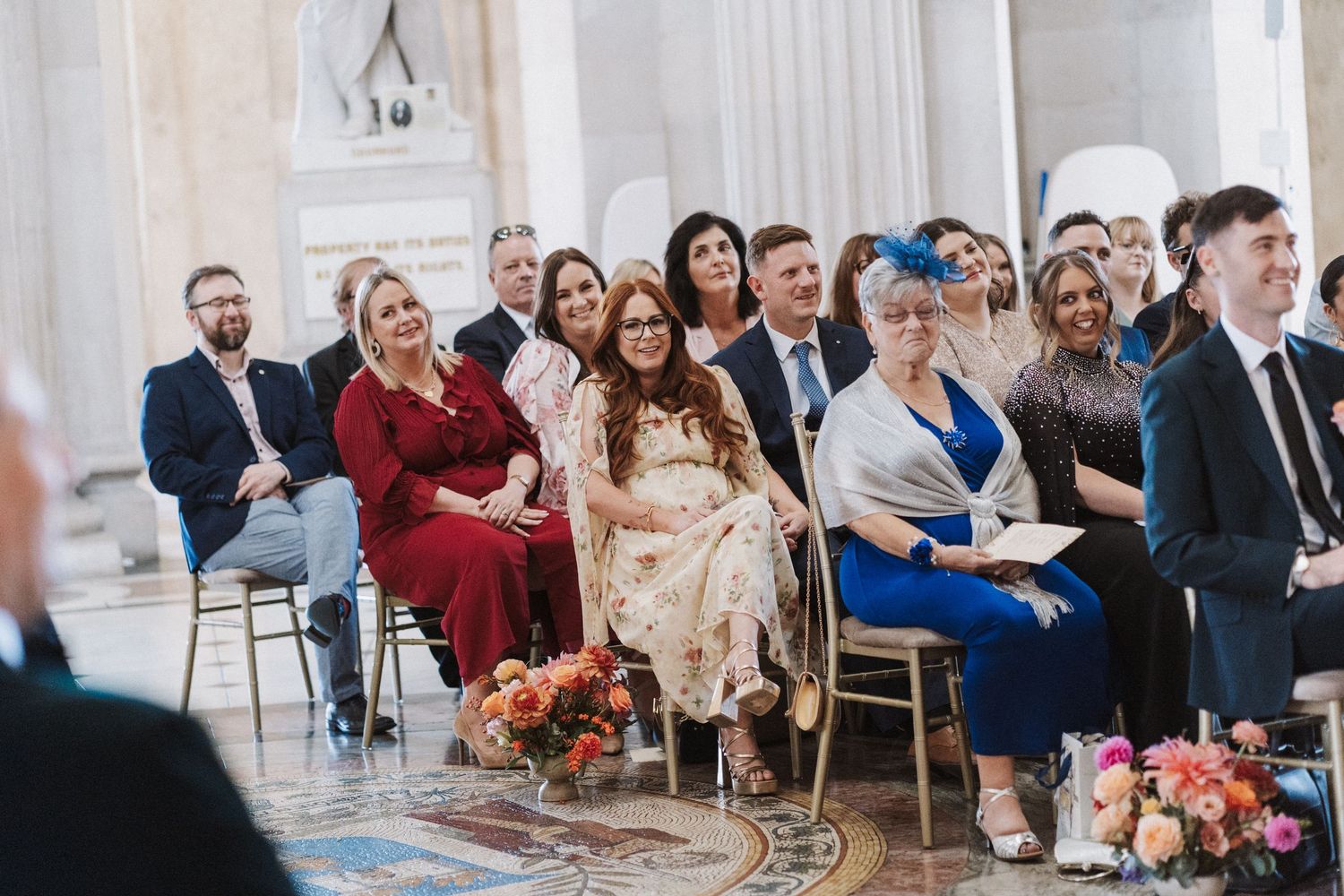 Wedding guests seated in elegant chairs watching a ceremony in an ornate venue with floral decorations.