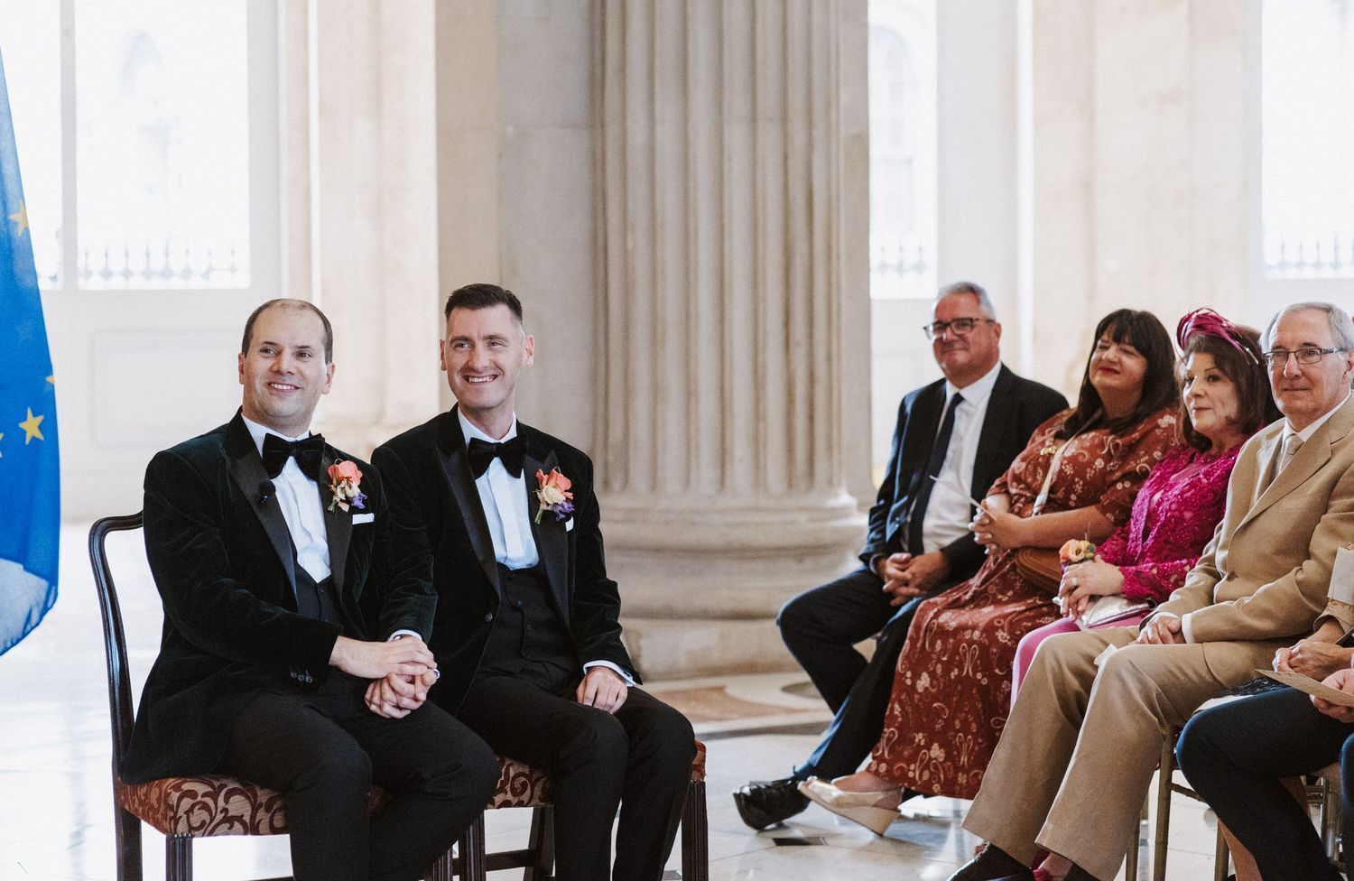 Wedding guests seated in elegant columned venue with formal decor and natural lighting.