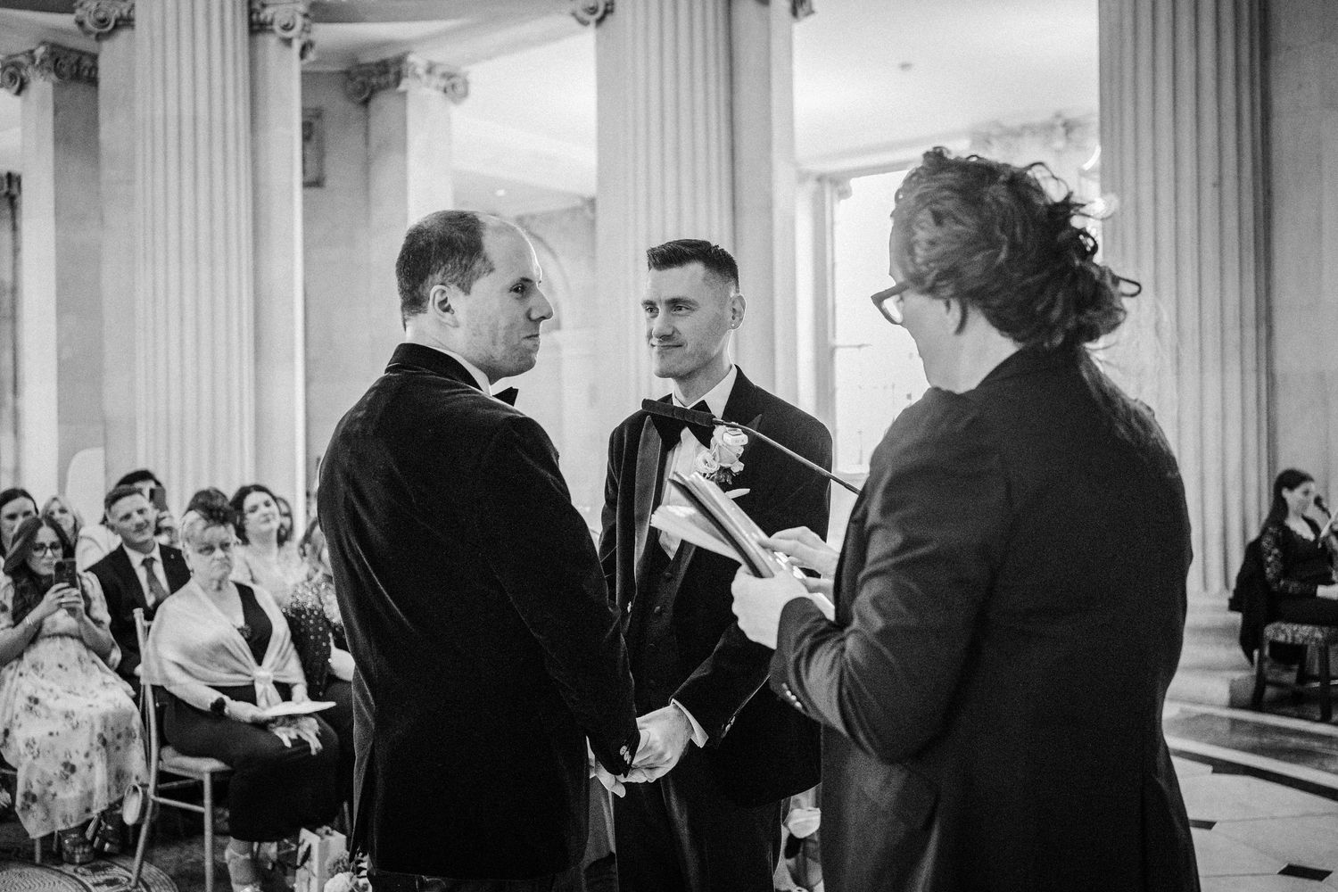 Black and white photo of wedding guests mingling in an ornate columned hall.