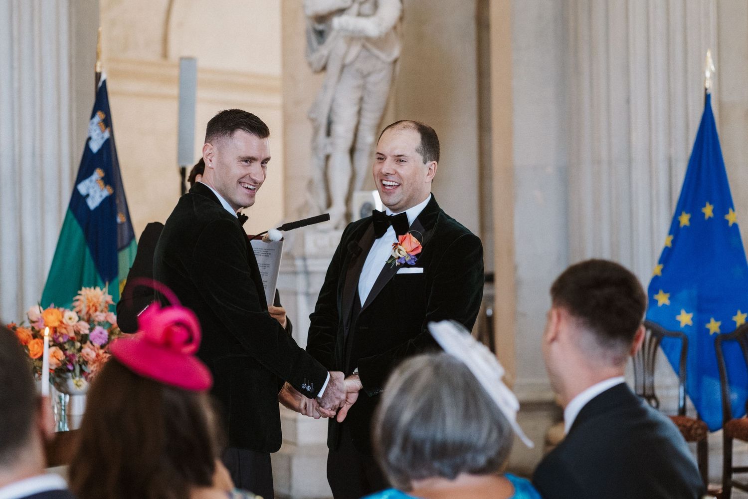 Formal wedding ceremony venue with classical columns and European flags in background.