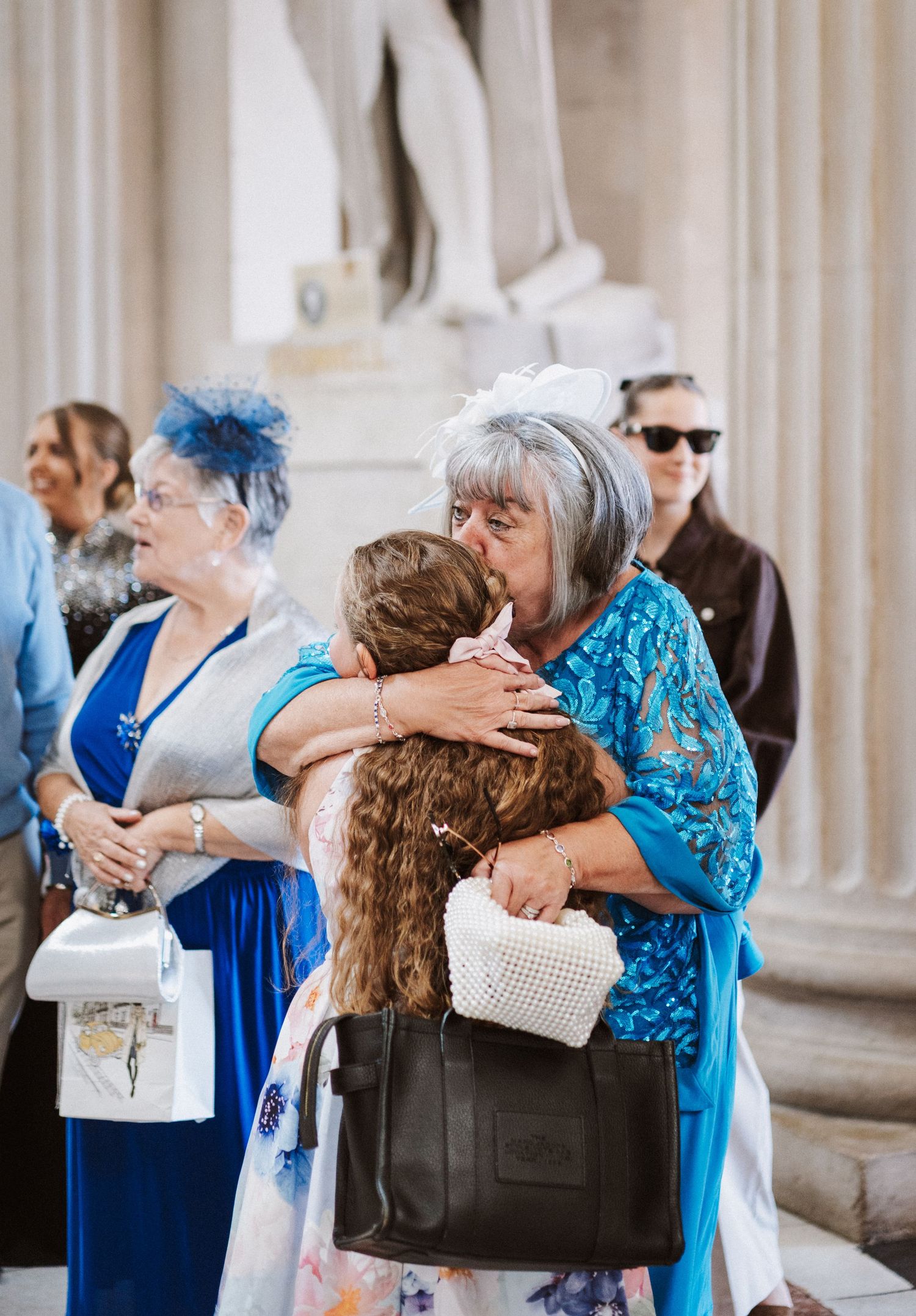 Wedding guests in blue and white attire embrace warmly in an elegant columned venue.