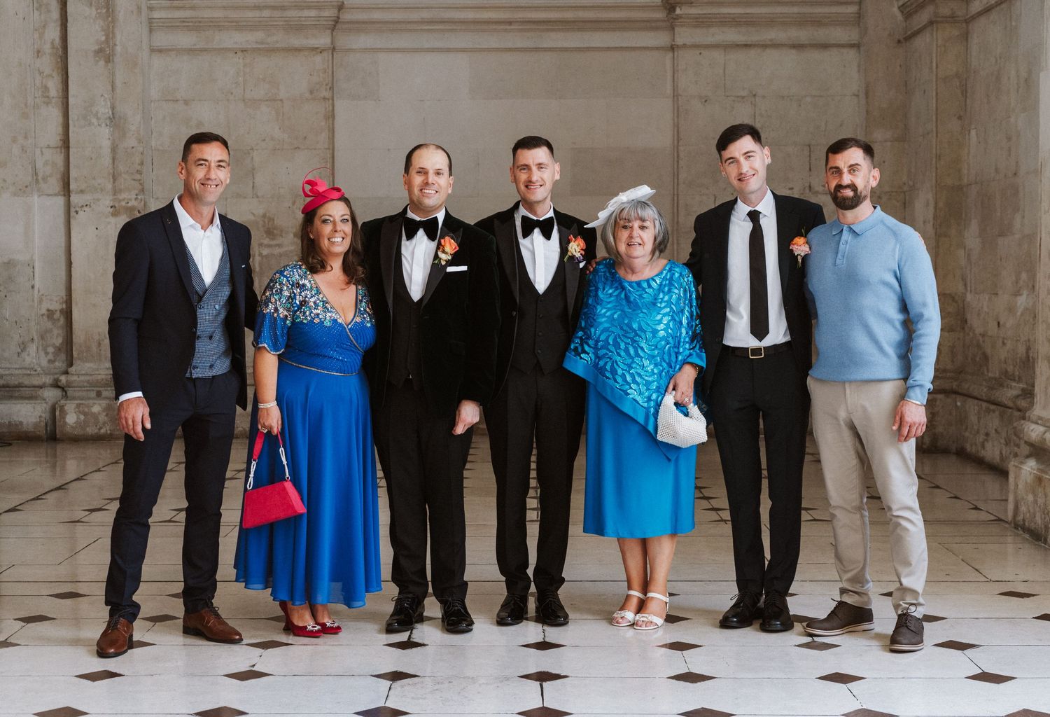 Formal group photo of wedding party members in suits and formal attire standing against a classic architectural backdrop.