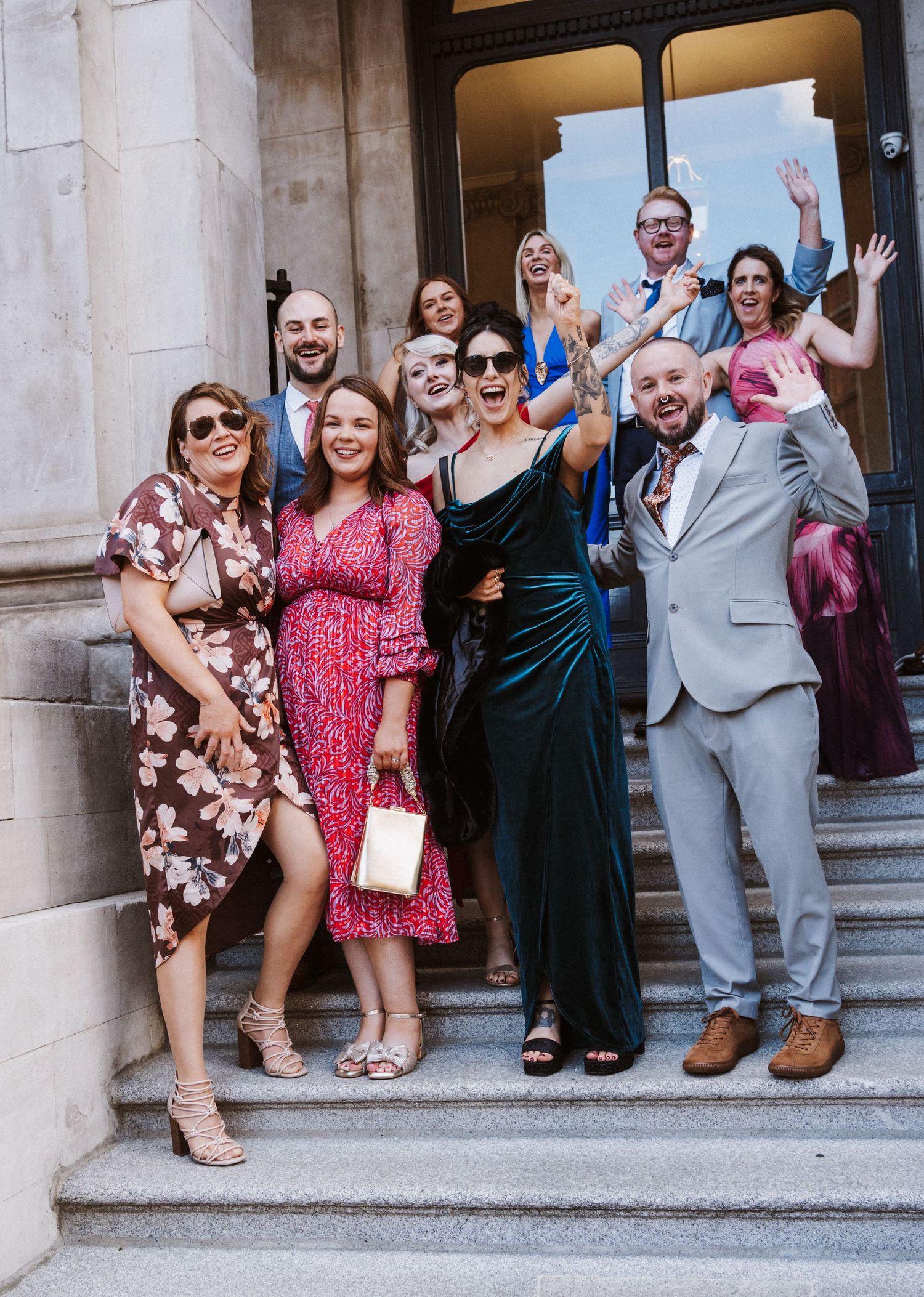 A group of wedding guests sitting and posing playfully on stone steps outside a building.