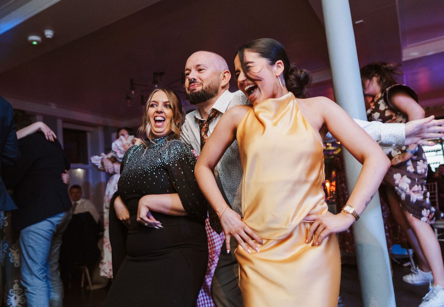 Joyful wedding celebration with guests dancing and laughing in a reception hall with purple lighting.