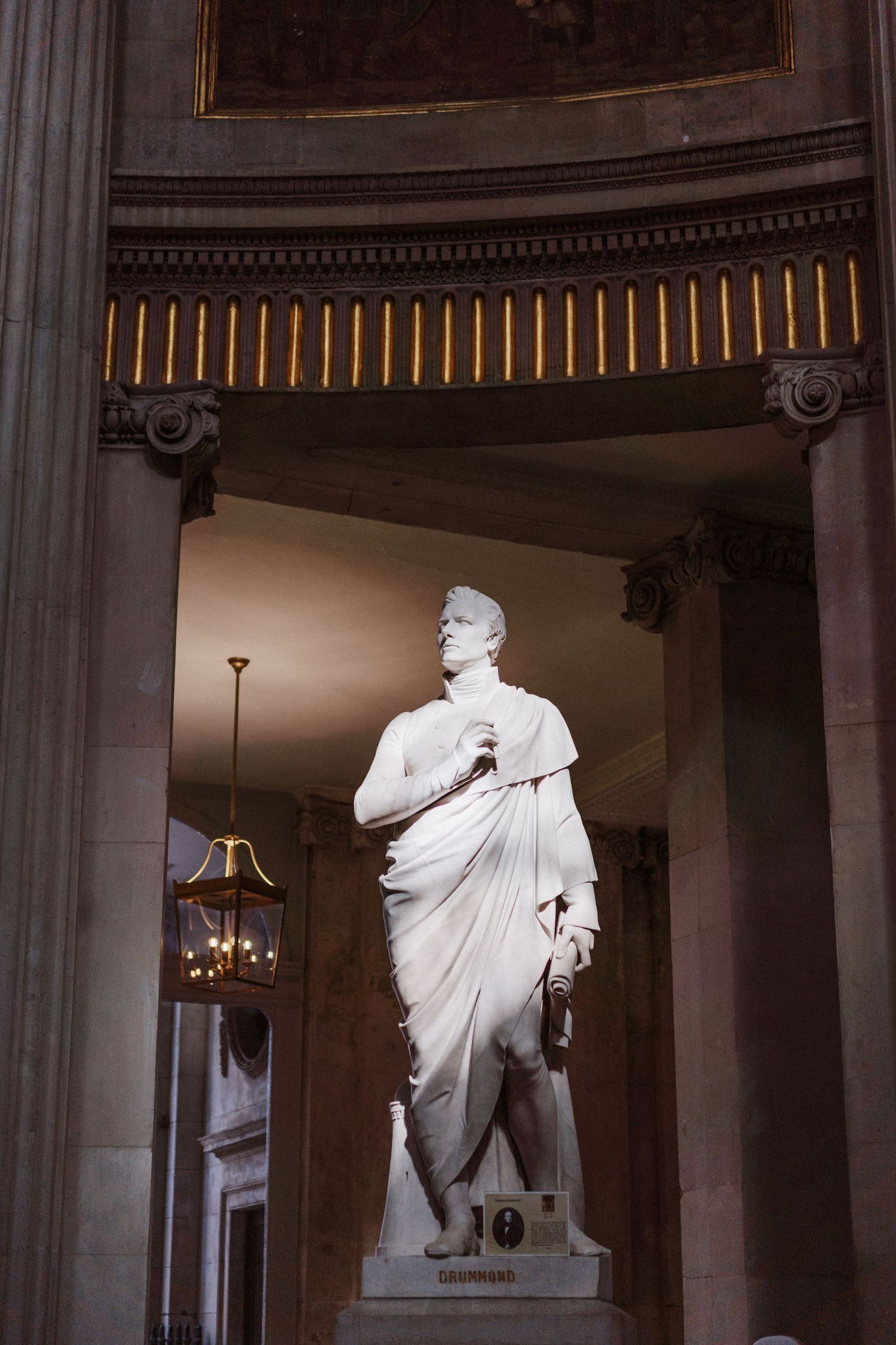 Classical white marble statue displayed in an ornate architectural alcove with decorative columns and overhead lighting.