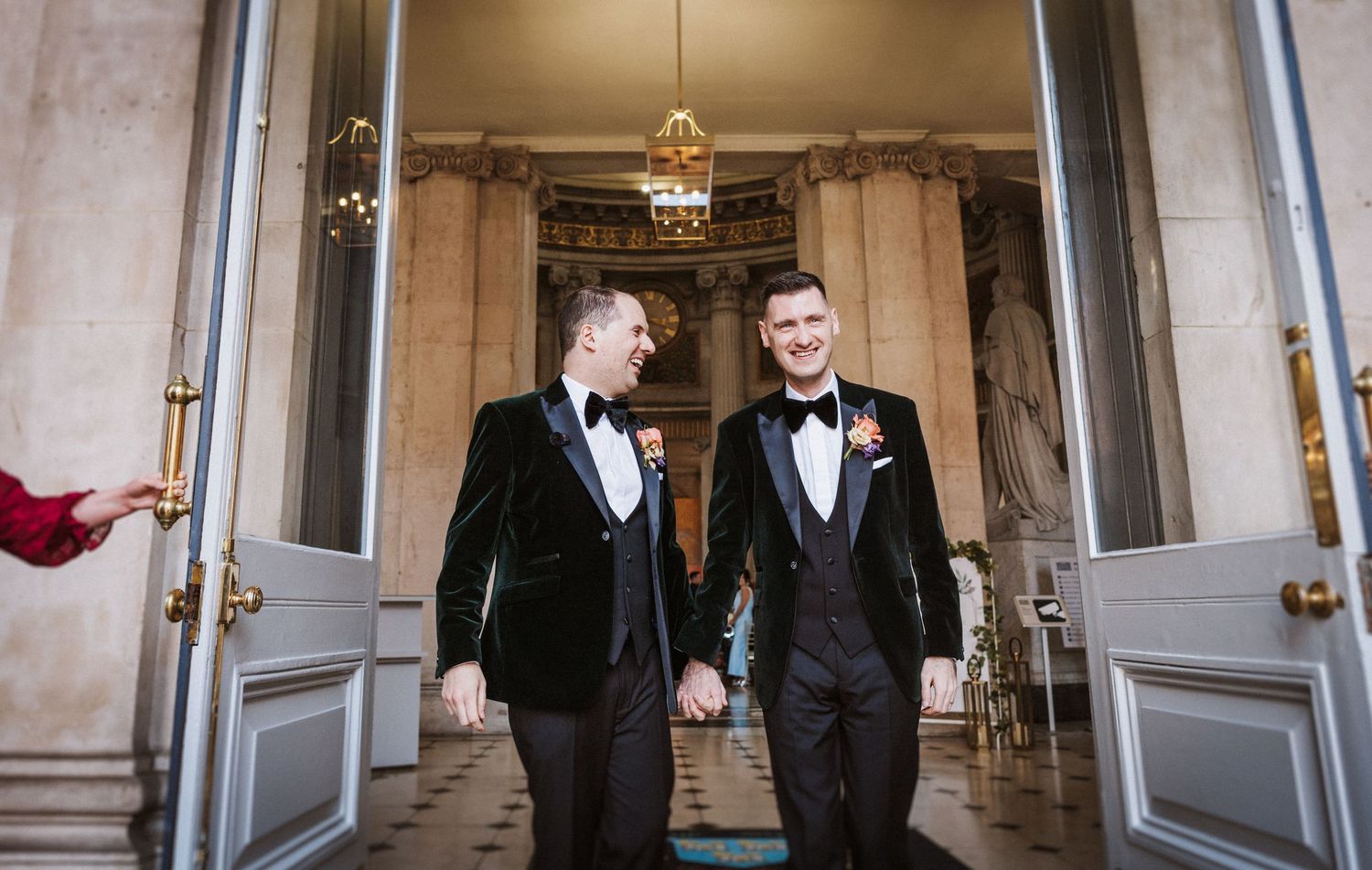 Two people in formal black tuxedos walk down an elegant marble hallway with ornate architectural details.