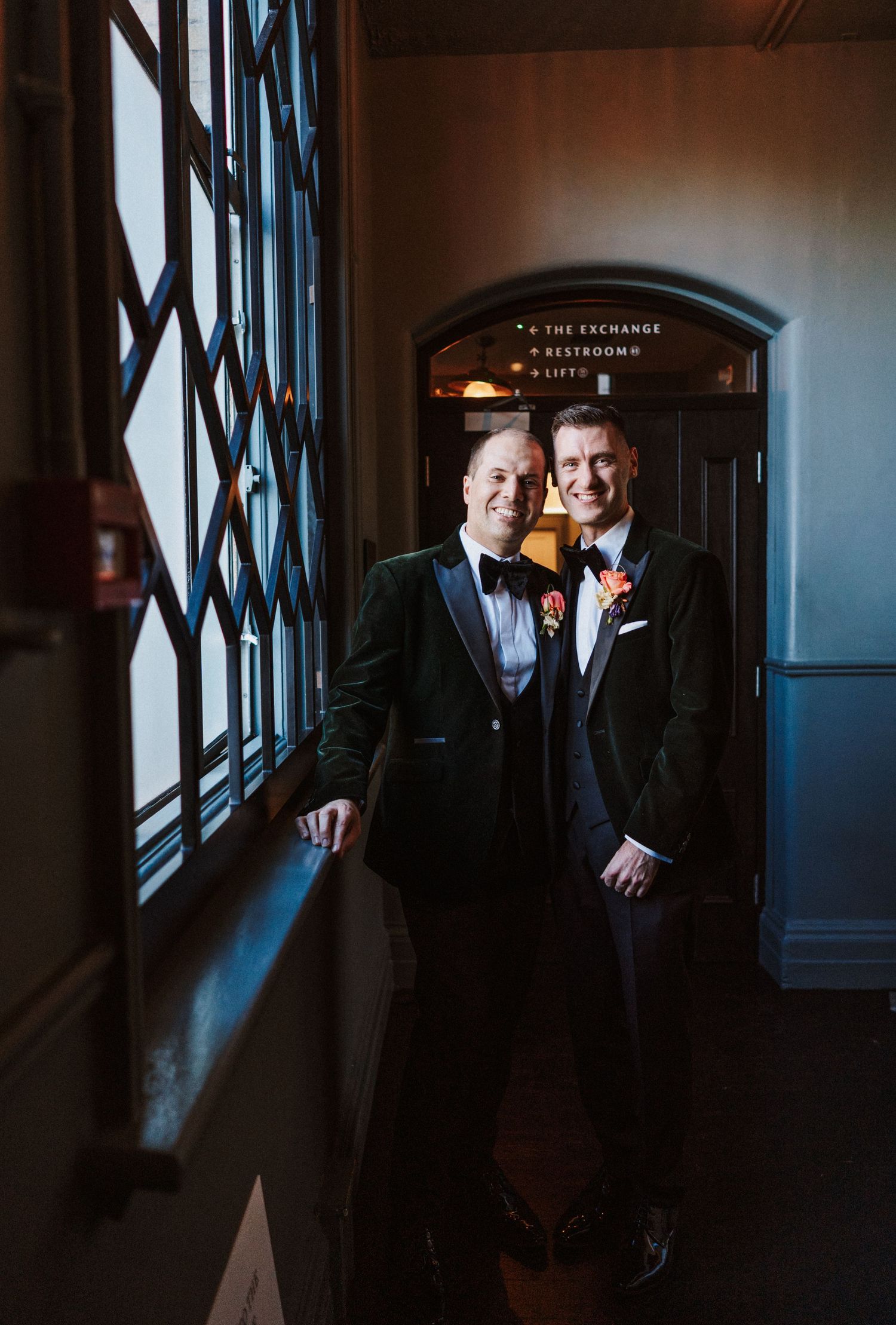 Two people in dark suits share an intimate moment in a dimly lit hallway with dramatic window lighting.