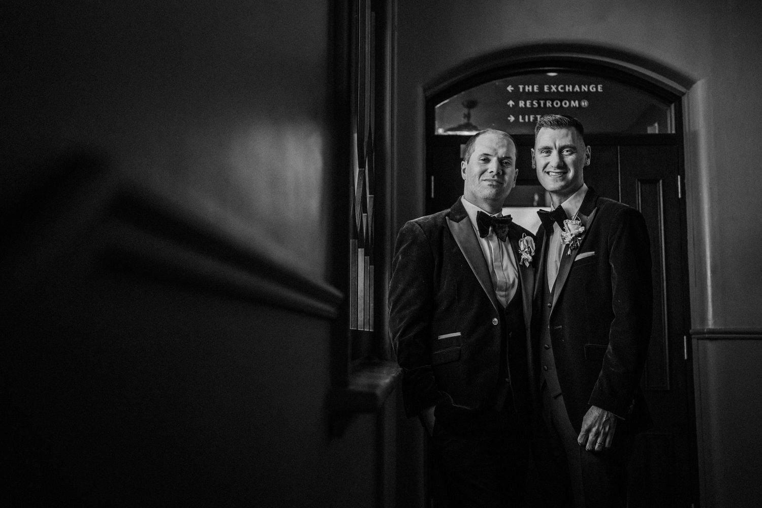 Black and white portrait photograph in dark dramatic lighting showing a person in formal attire against a shadowy background.