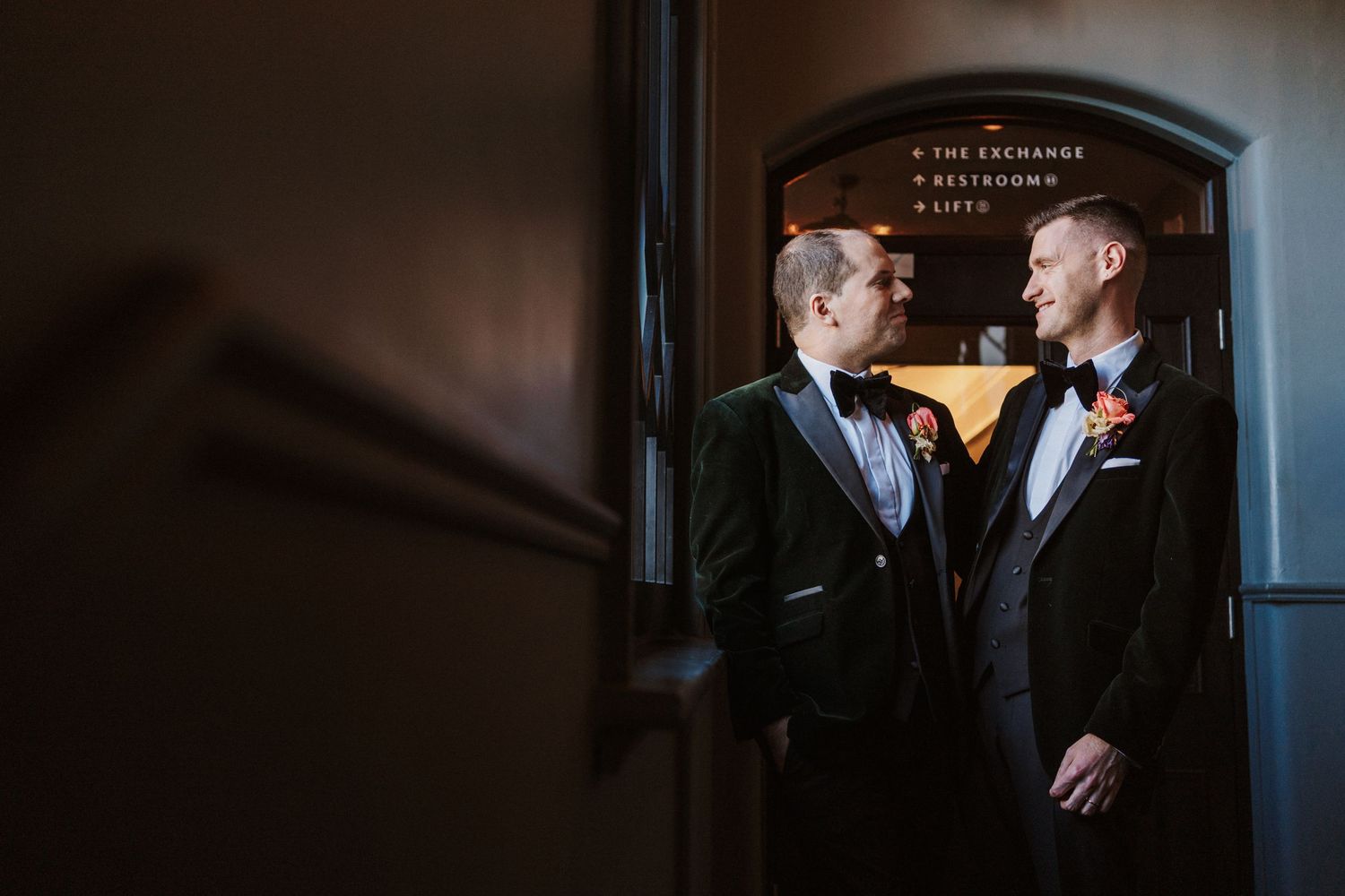 A couple in formal wedding attire share an intimate moment in a dimly lit hallway.