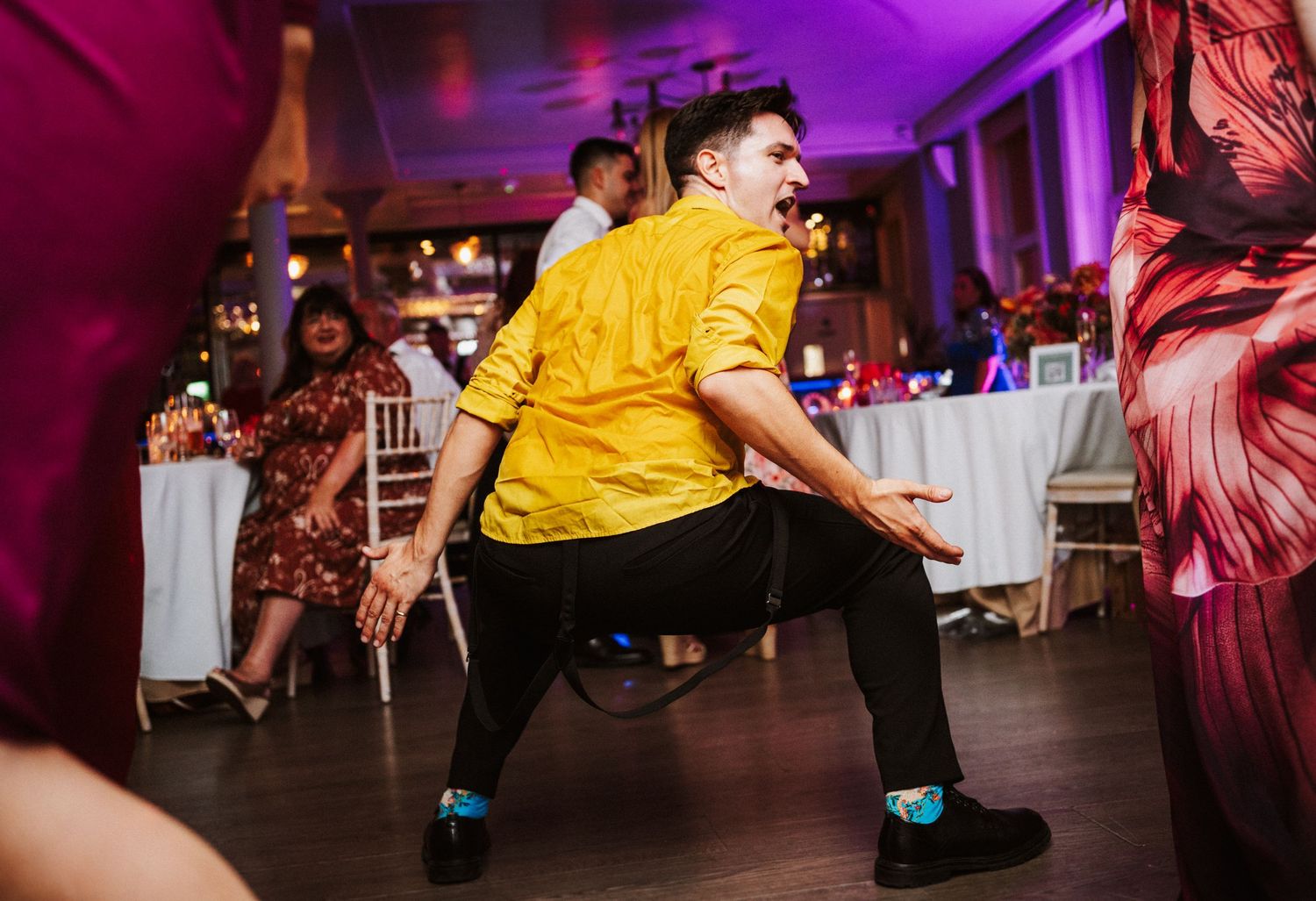 People dancing and celebrating on a dance floor illuminated by vibrant purple and pink lighting at an indoor event.