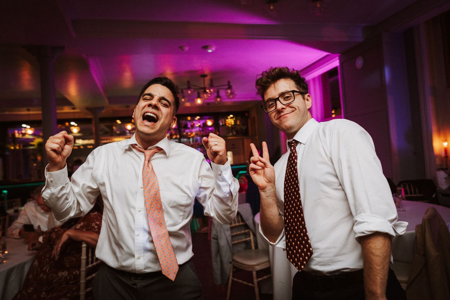 Two people in white dress shirts and ties dance energetically at a reception with purple lighting in the background.