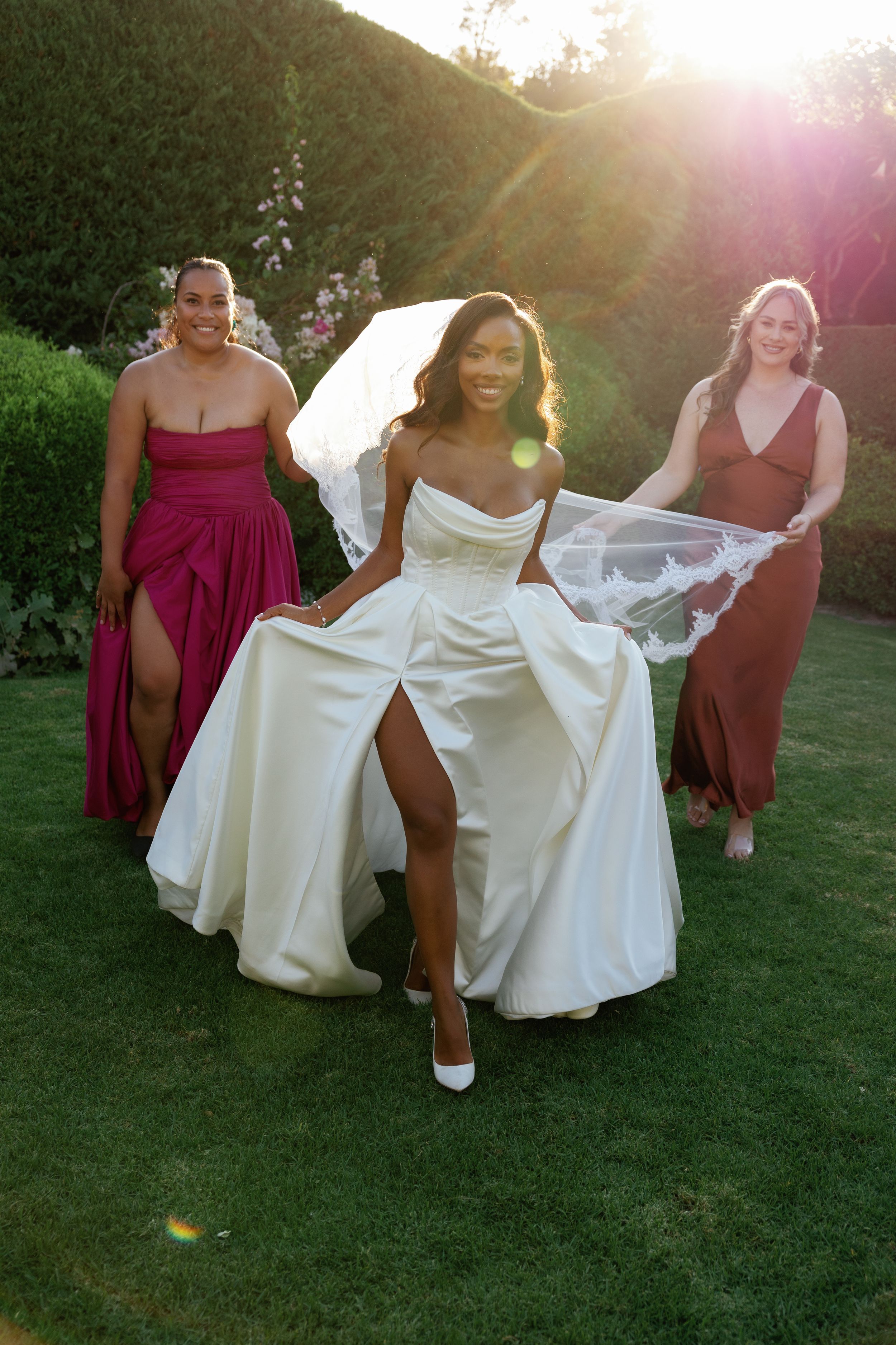 Bridesmaids in burgundy dresses dance and celebrate on a grassy lawn at sunset during a wedding celebration.