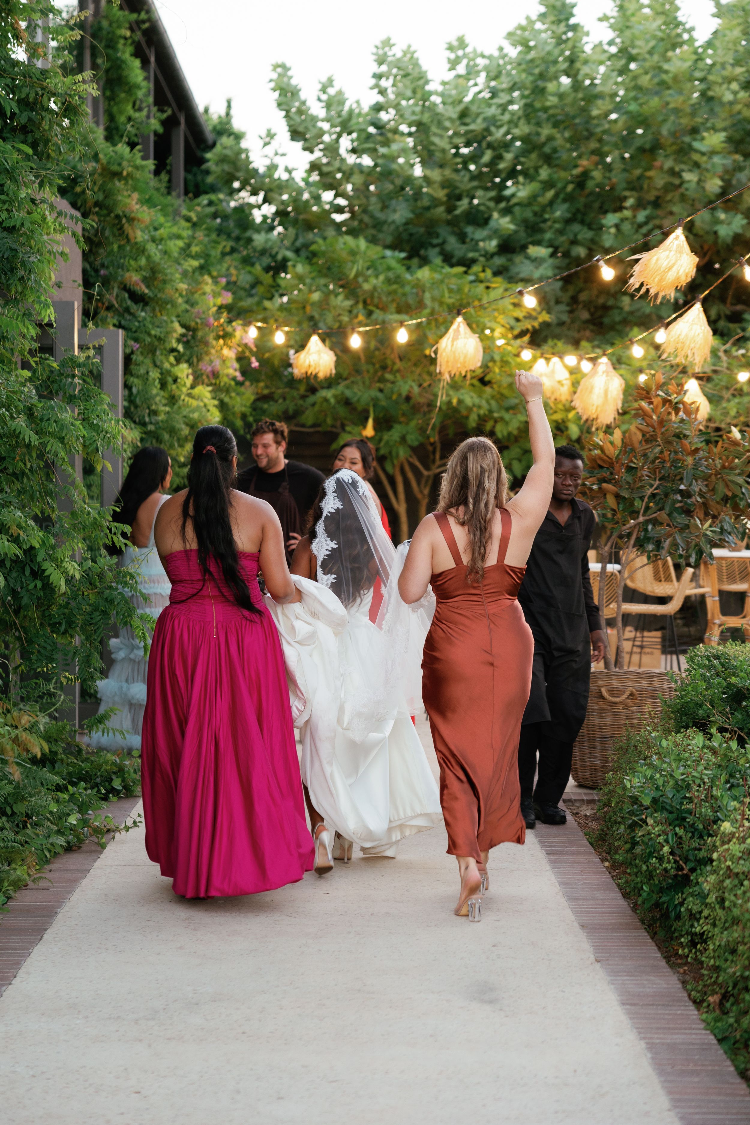 Wedding party walks together on garden path with string lights overhead during golden hour celebration.