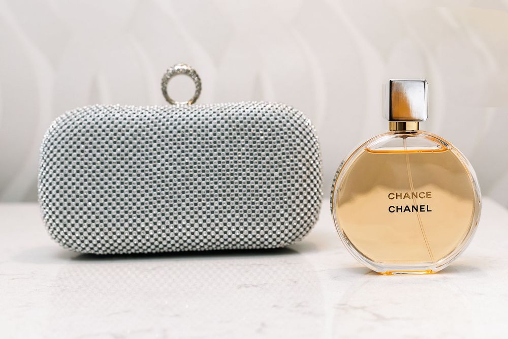 A silver rhinestone evening clutch purse sits next to a bottle of Chanel Chance perfume on a white surface.