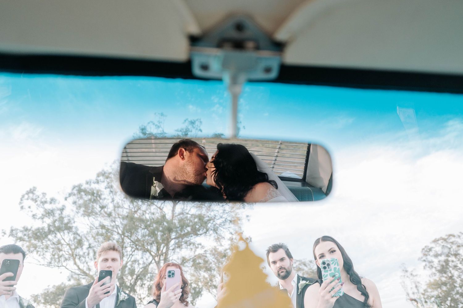 View through car rearview mirror showing reflection against bright blue sky and trees in background.