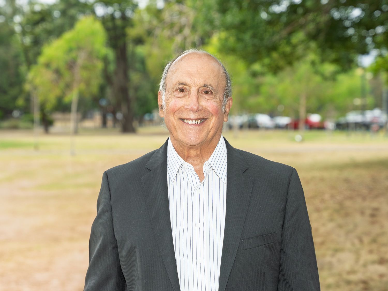 Smiling senior businessman in a blazer stands outdoors in a sunny park with green trees behind him.