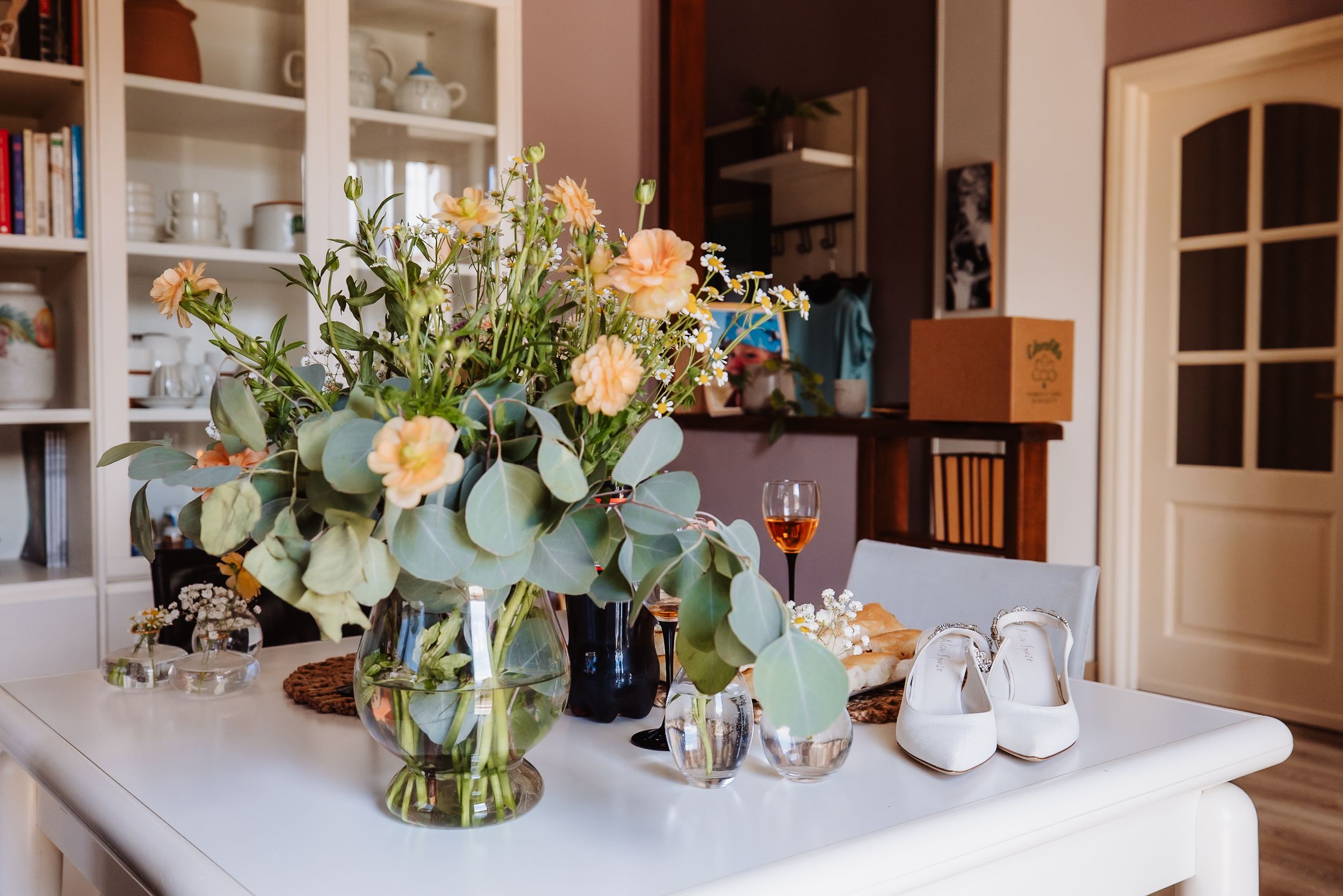 A beautiful floral arrangement with peach roses and eucalyptus leaves sits on a white table next to wedding shoes.