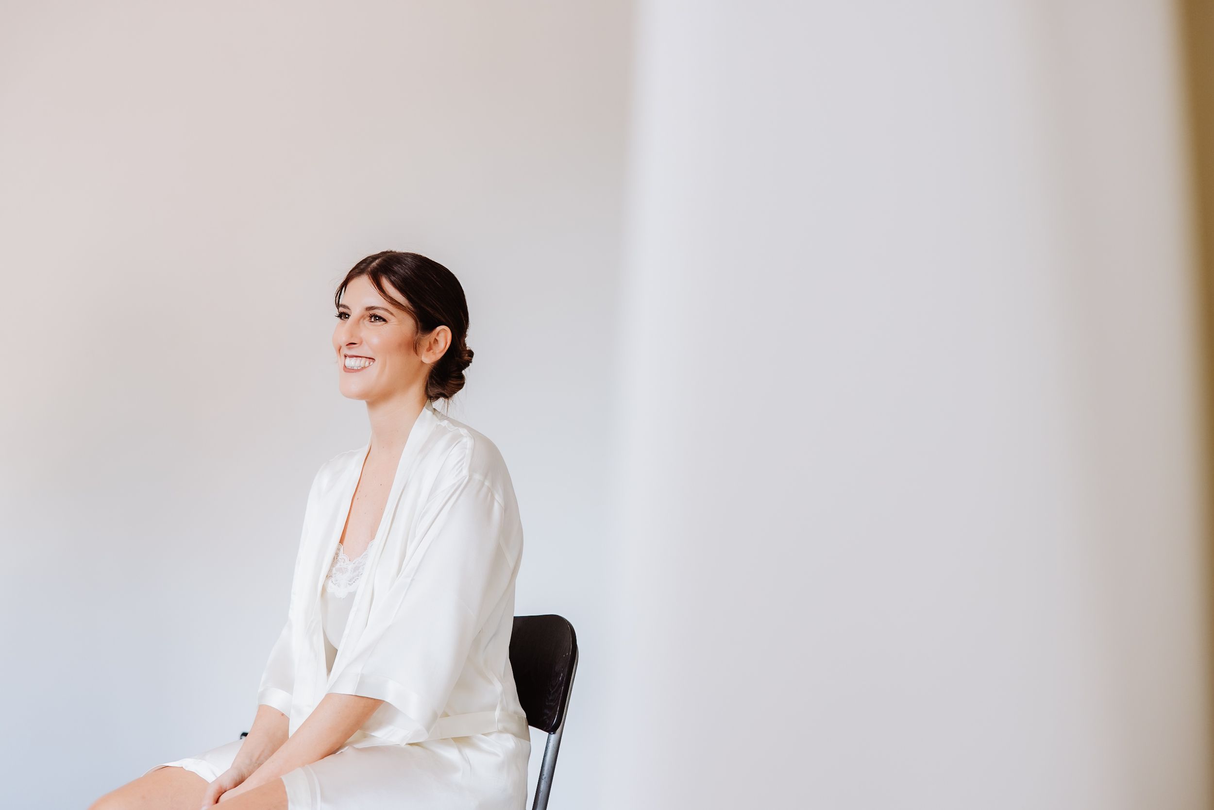 A woman in a white blouse and dark pants smiles while sitting on a chair against a light-colored wall.