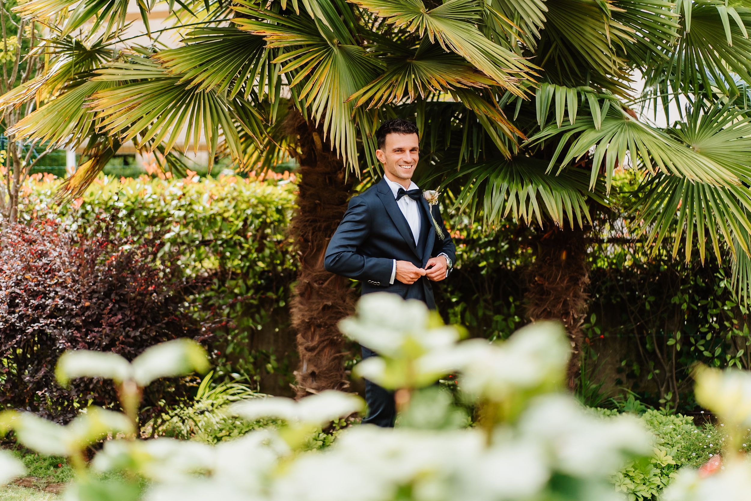 A groom in a classic black tuxedo stands among lush tropical palm trees and garden foliage.