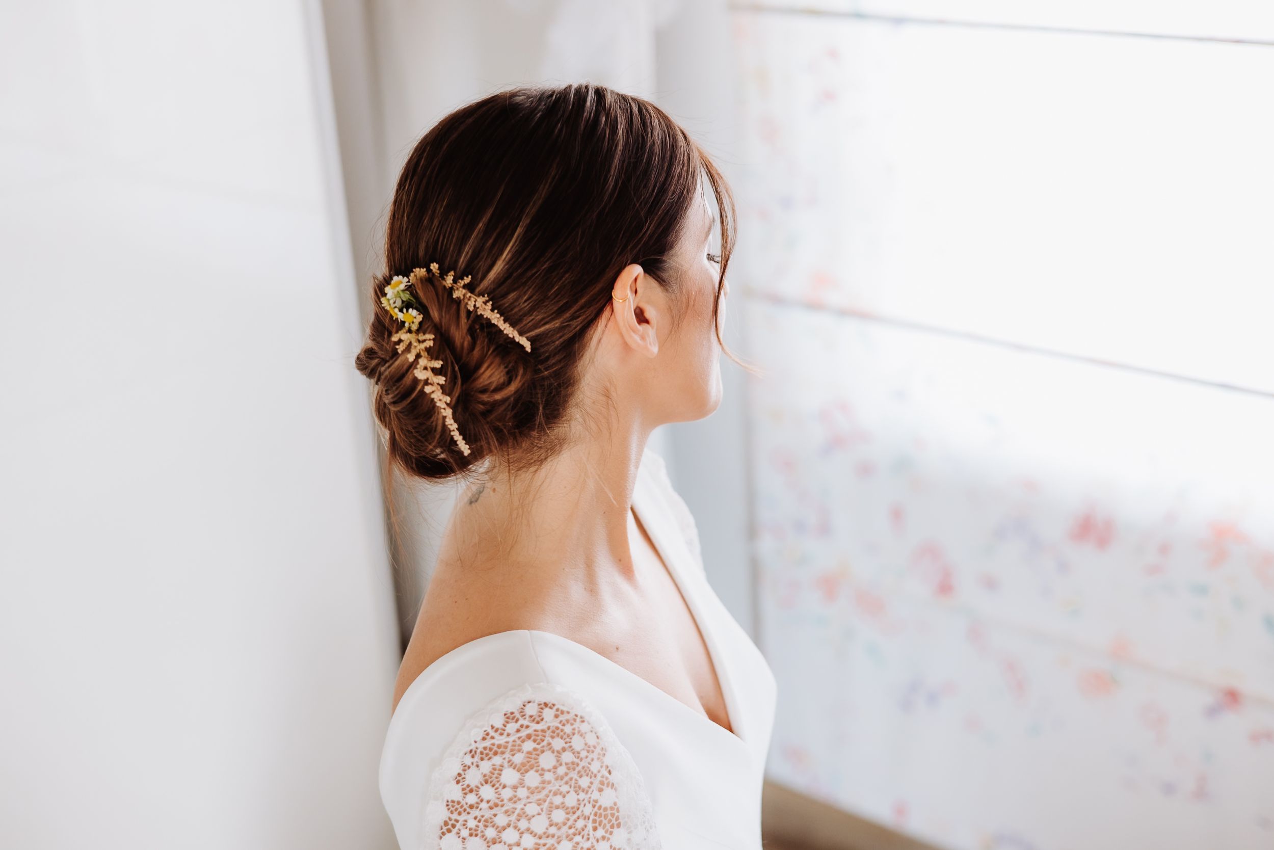 A bride in a delicate lace wedding dress with a romantic updo hairstyle stands by a bright window.