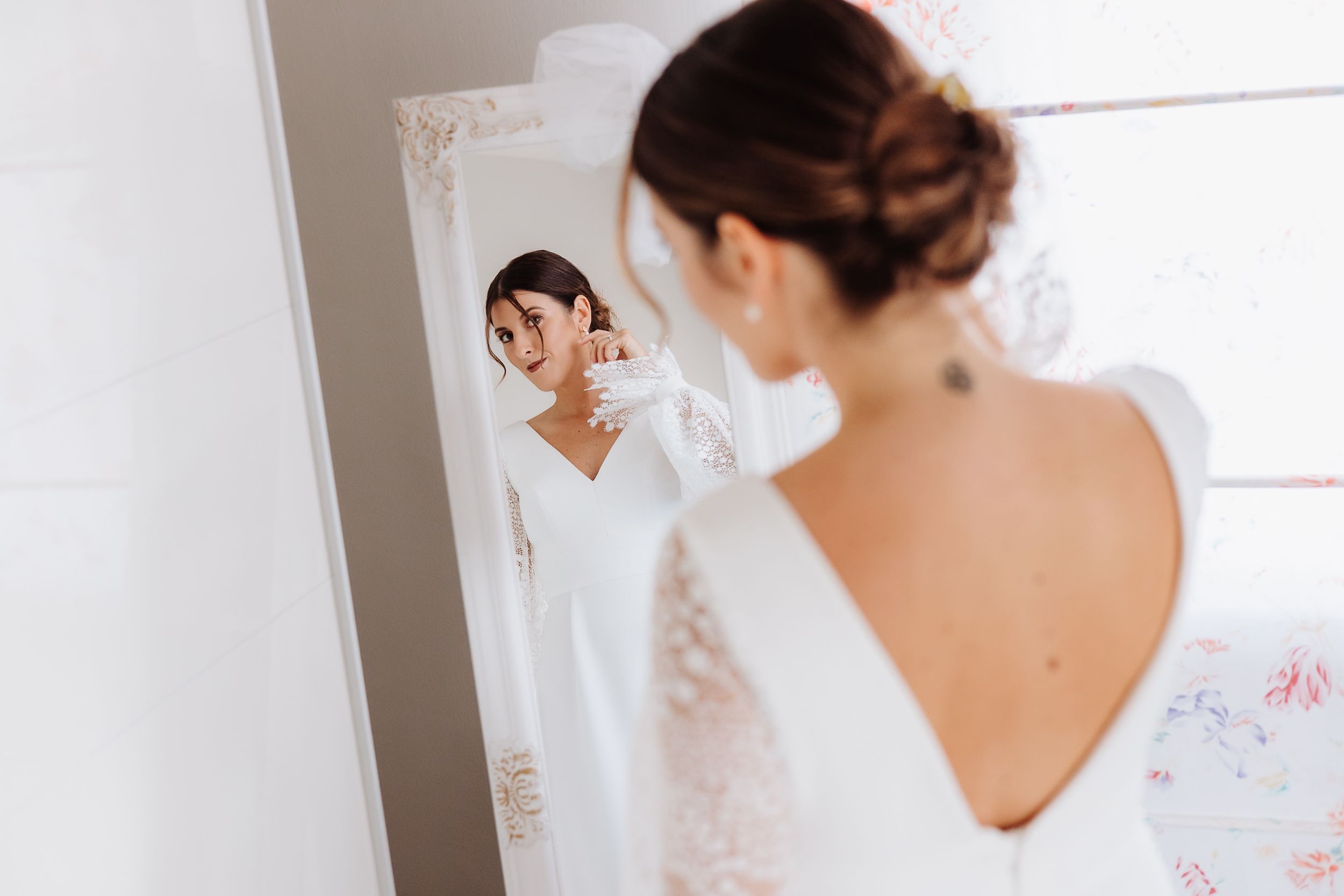 A person in white bridal attire looks at wedding dresses hanging in a bright, sunlit room.