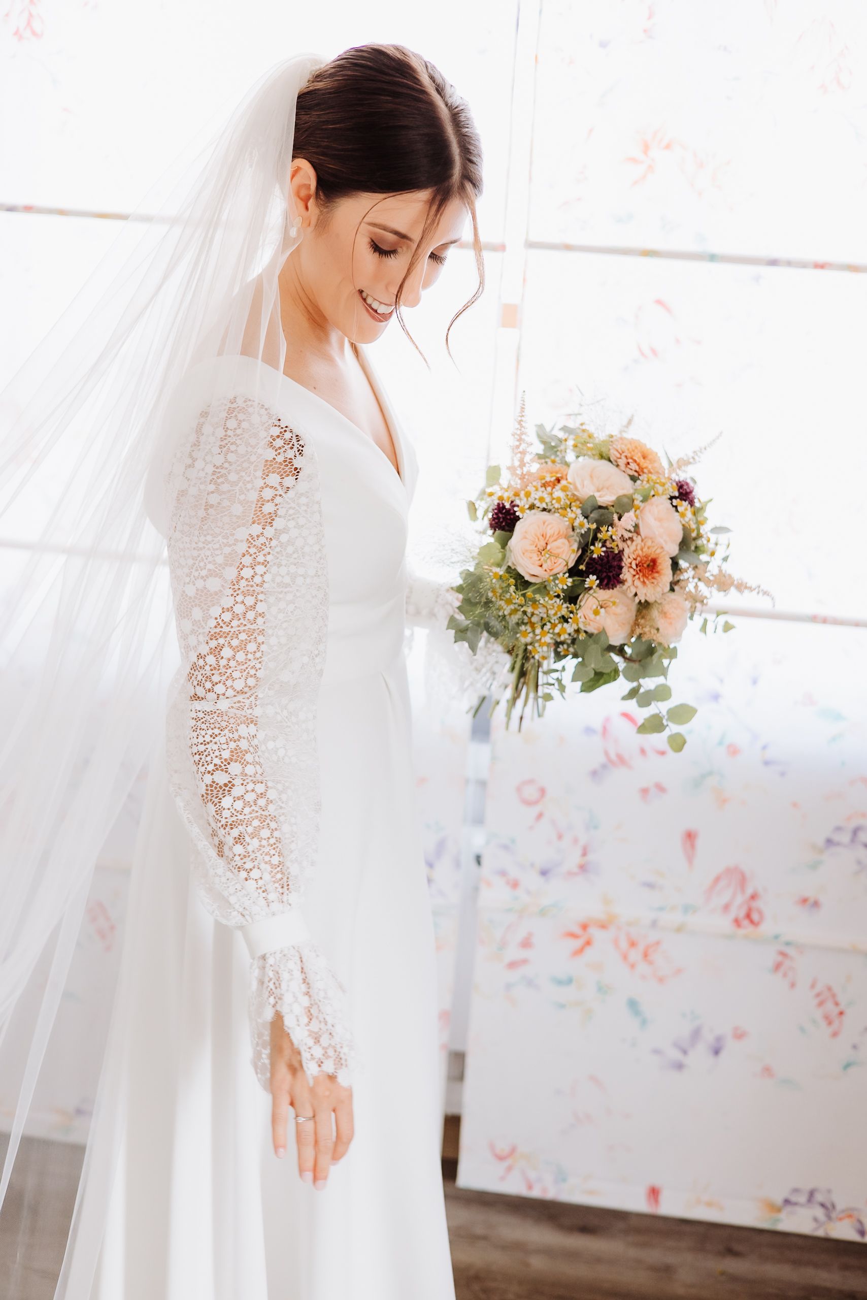 A bride in a white lace-sleeved wedding dress holds a peach and blush floral bouquet against a light backdrop.